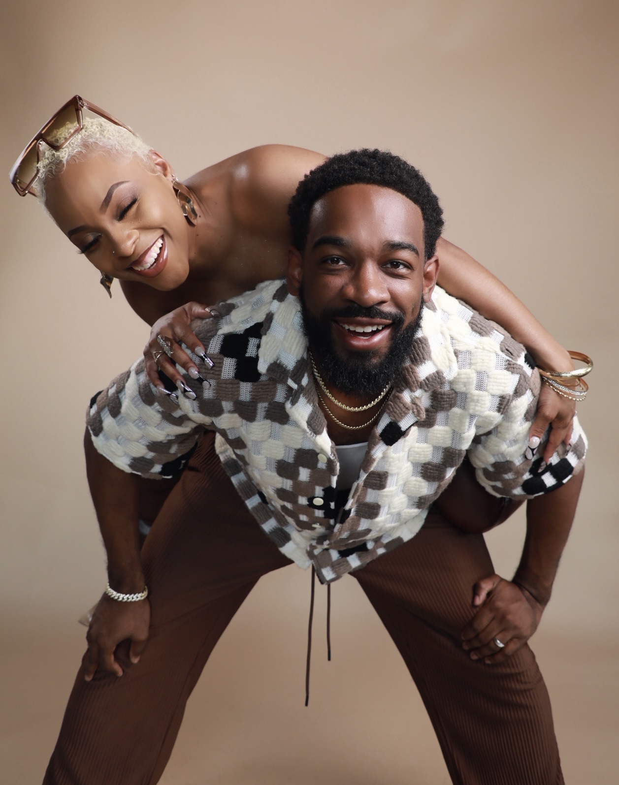 A smiling woman with platinum blonde hair and a man with a beard pose together, woman leaning on man's back, both smiling.