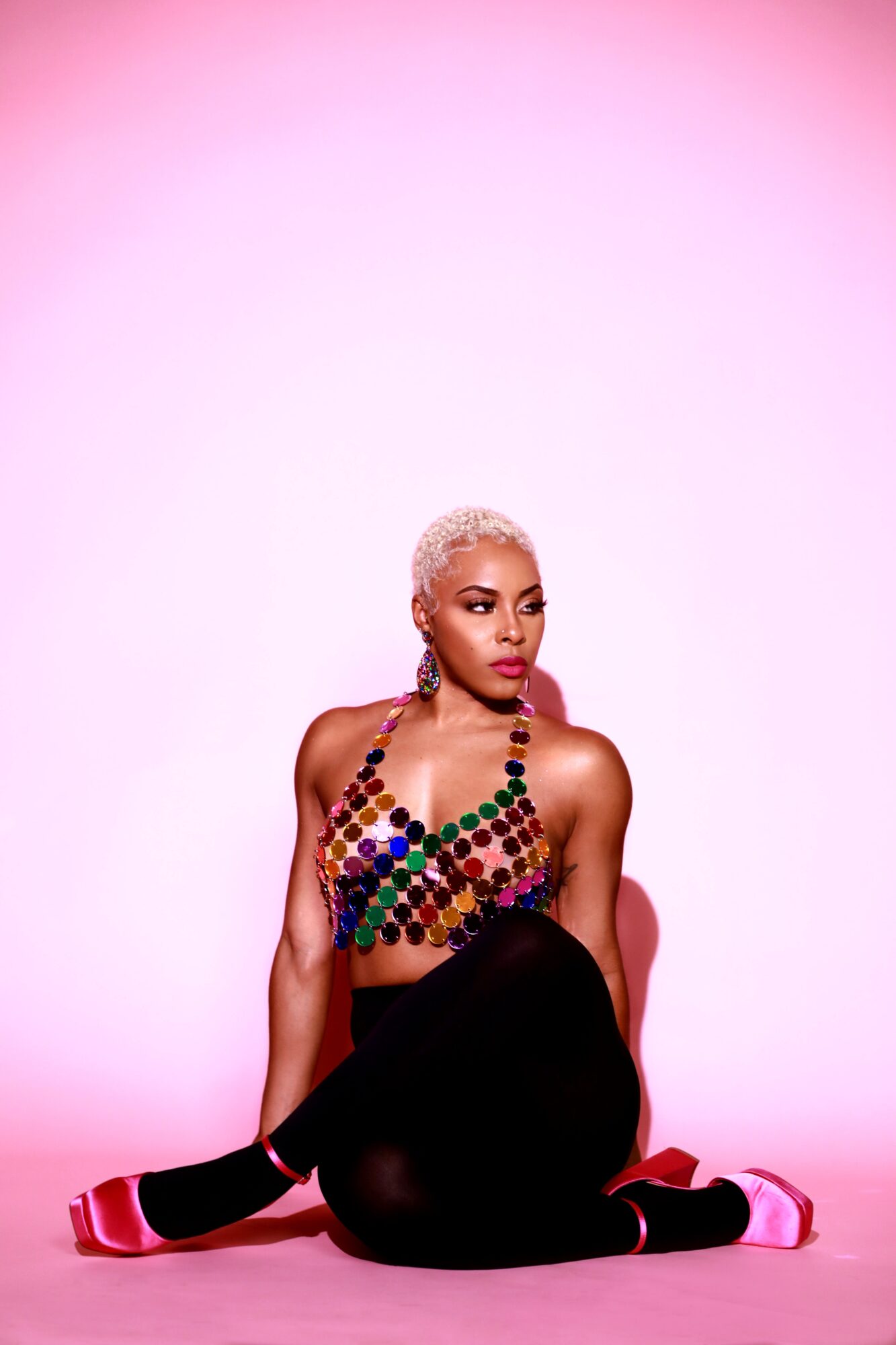 Person with short platinum hair sitting against a pink background, wearing a colorful beaded top and black pants.