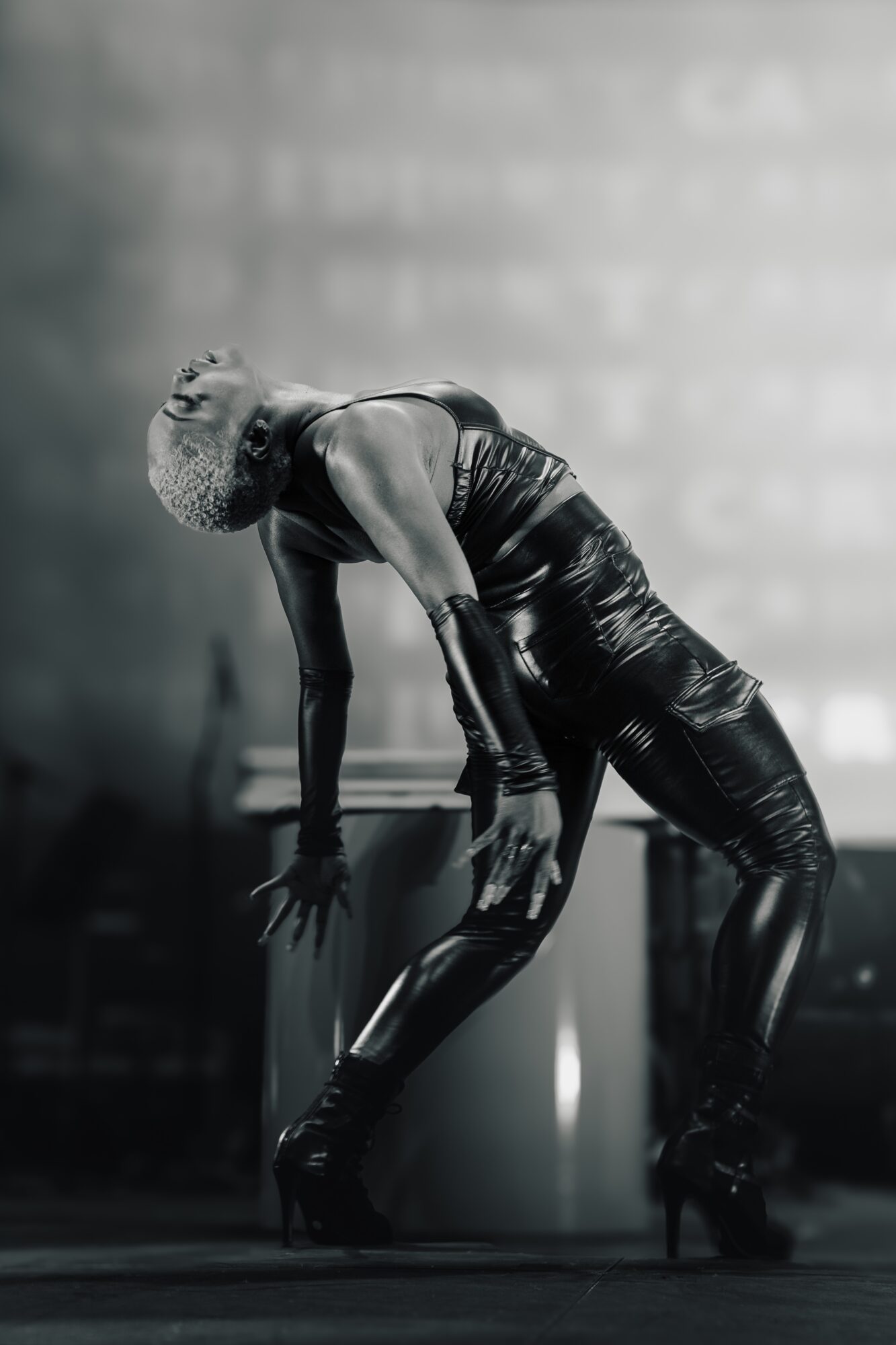Performer with curly hair bent forward, hands on knees, on stage in black outfit, in black and white photo.