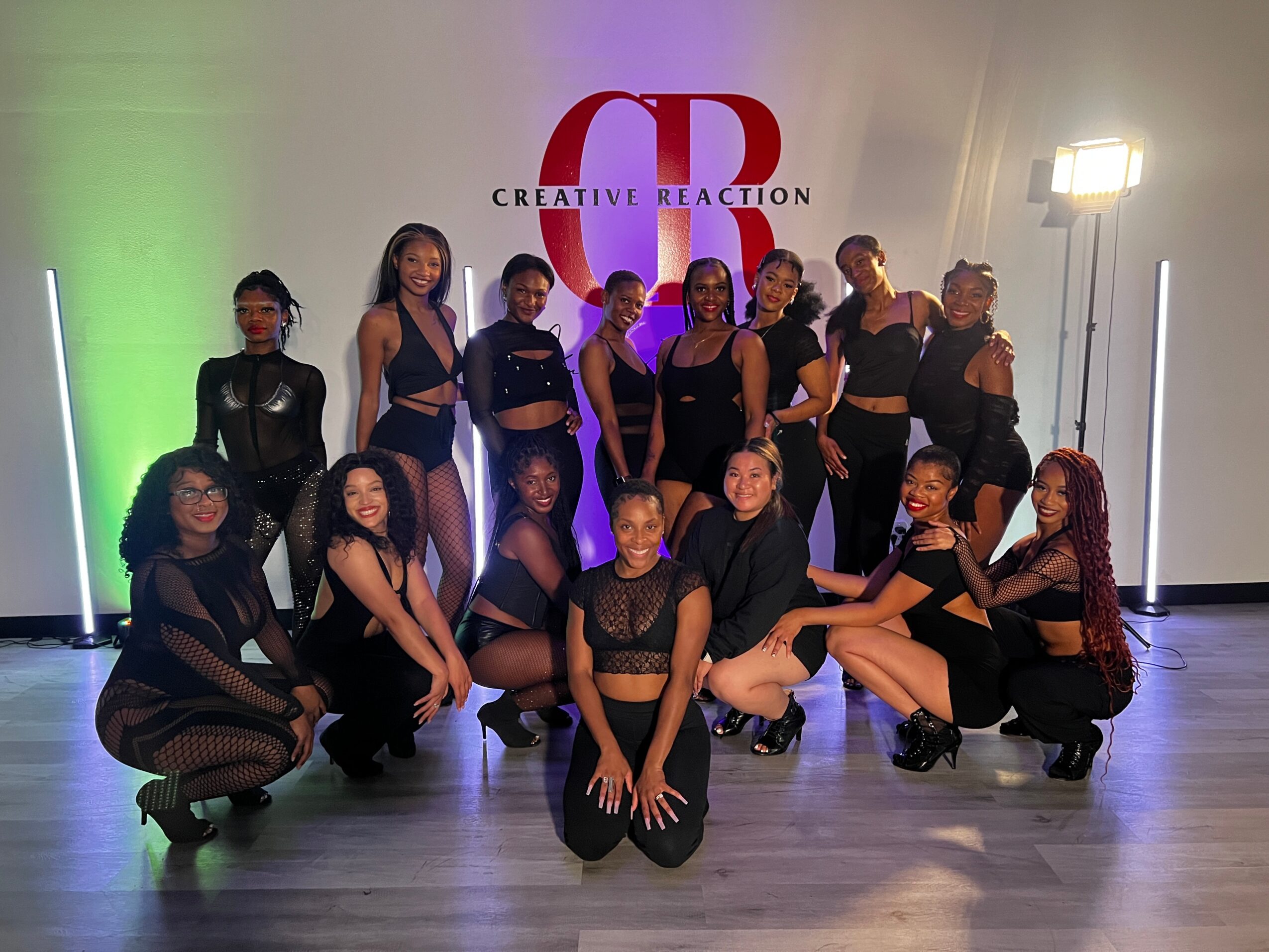 Group of 14 women in black outfits posing in front of a wall with a red and black logo, some kneeling and some standing.