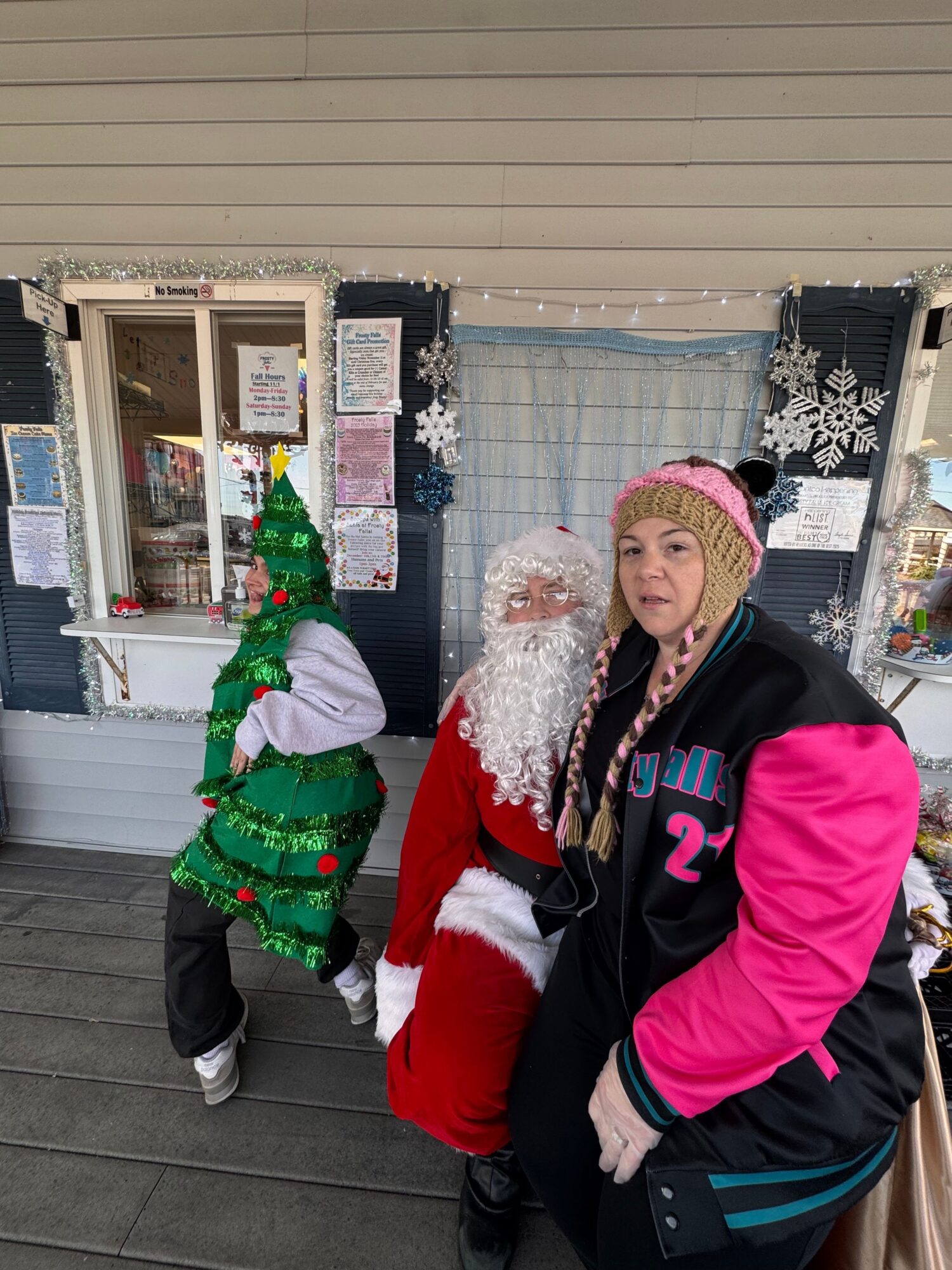 Woman in winter clothing and Santa costume with a child in a green Christmas sweater and hat outside a store.