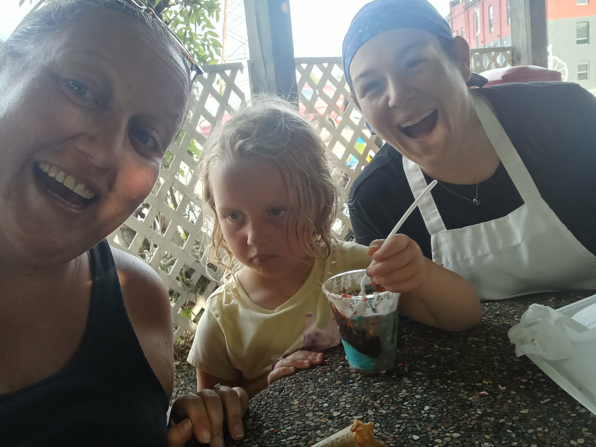 Three people smiling at a table outdoors, with a young girl in the middle, two adults on either side, and a drink in front.