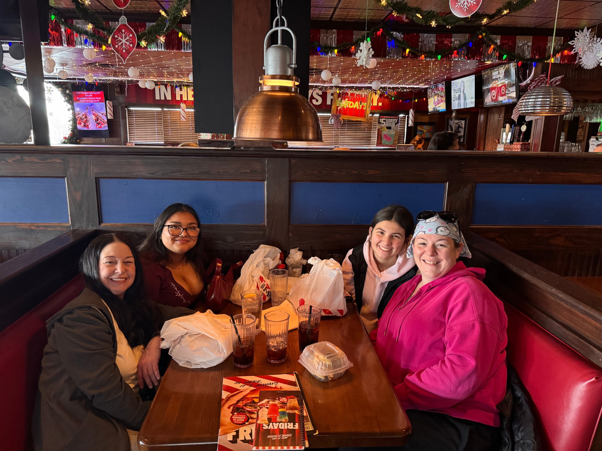 Four women sitting at a restaurant table with food and drinks, smiling, with a decorated interior background.