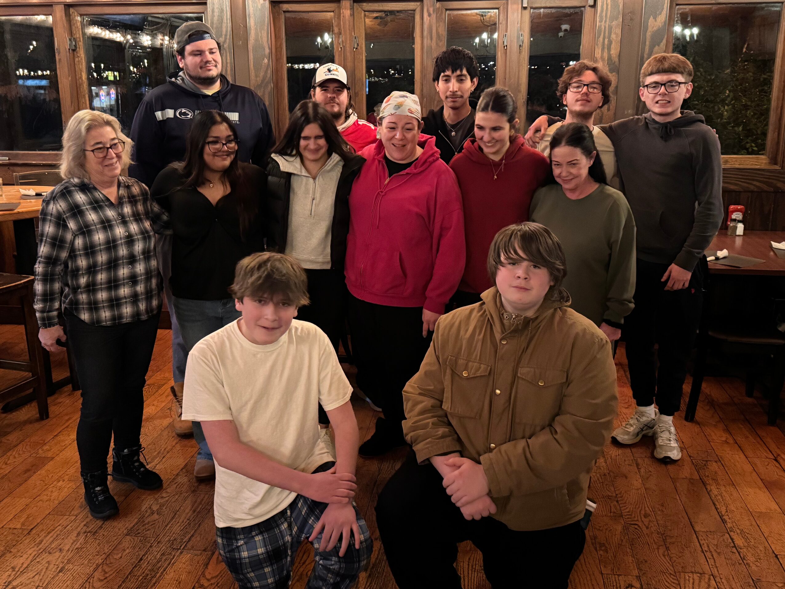 Group of fifteen people, some standing and some sitting, in a room with wooden floors and large windows, smiling at the camera.