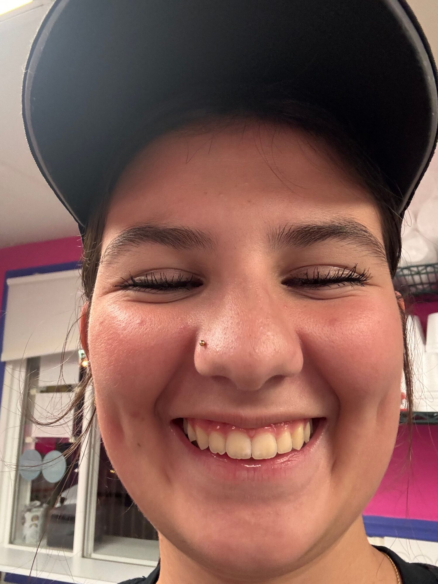 Close-up of smiling person with eyes closed, wearing a black cap, in a room with pink and white background.