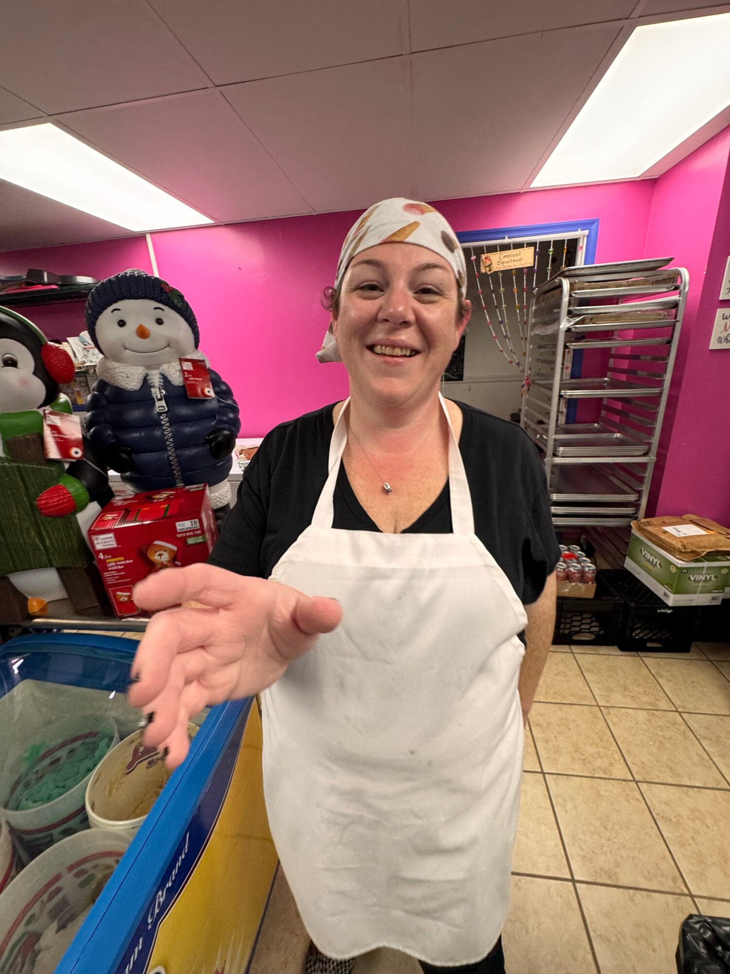 Smiling woman wearing a white headscarf and apron, standing in a colorful kitchen with pink walls, reaching out with her hand.