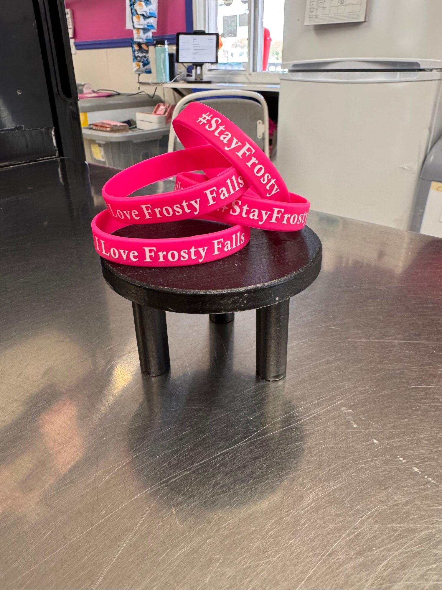 Cake with pink ribbon reading 'Stay Frosty' on a black stand, on a metallic surface.