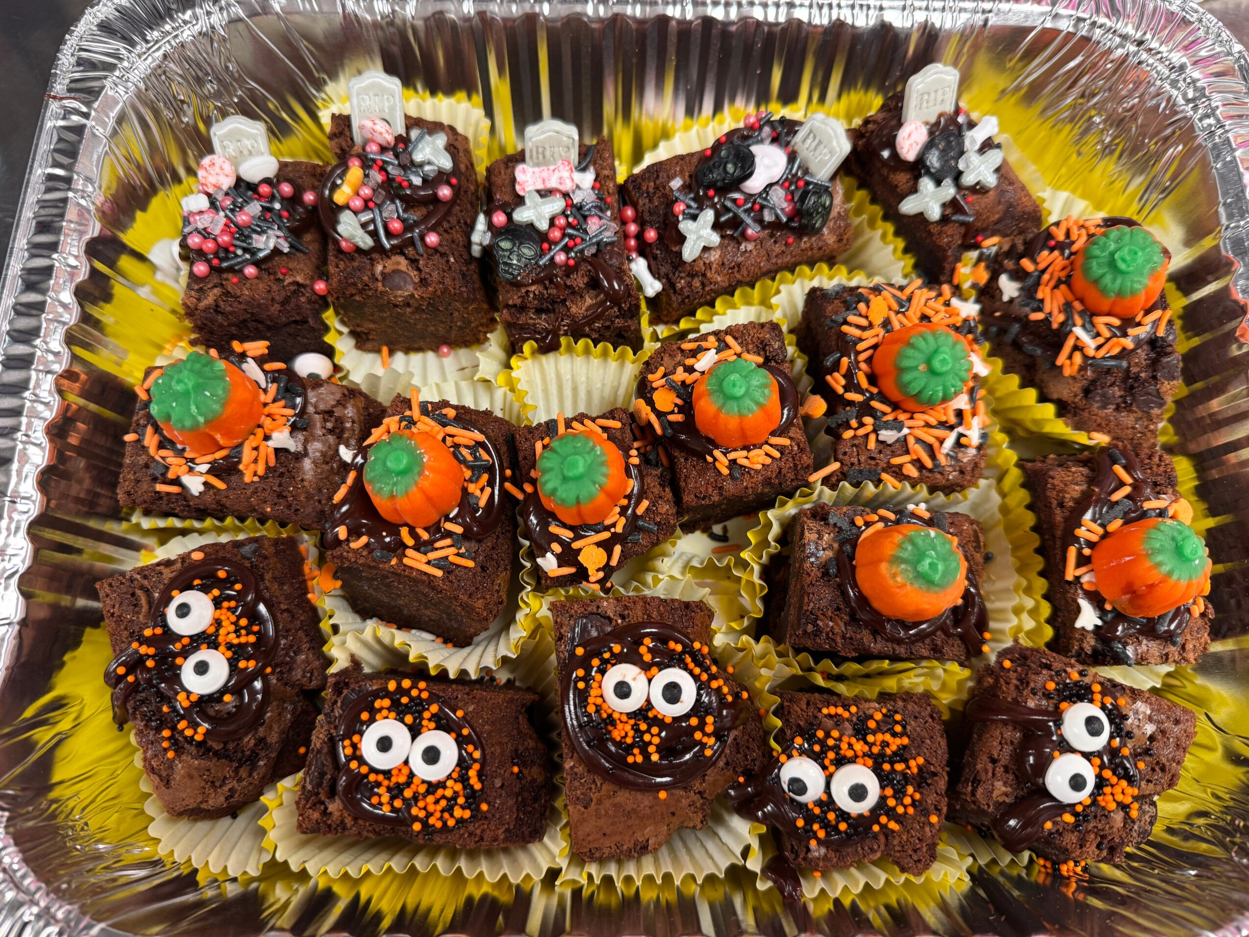Assorted decorated brownies with orange, green, and white toppings arranged in a tray, some with googly eyes, on yellow paper cups.