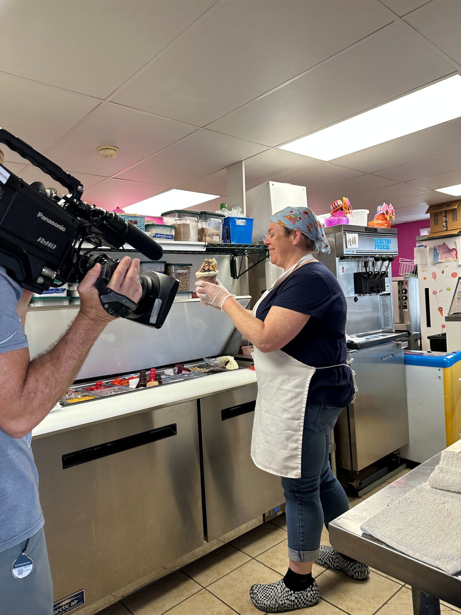 Person in a kitchen preparing food, wearing a blue shirt, apron, and hairnet, with a camera operator filming.