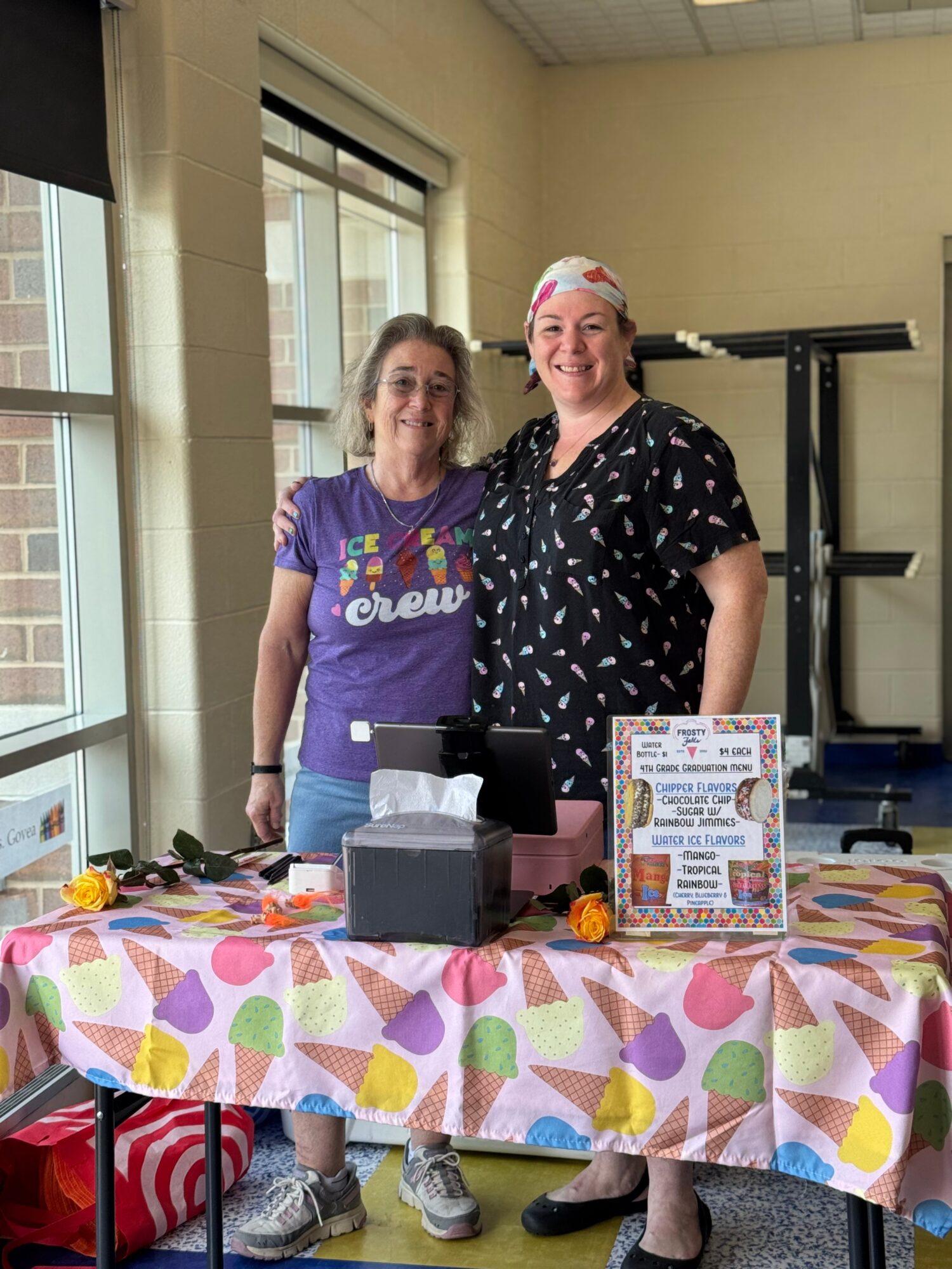 Two women standing behind a table with colorful tablecloth, smiling indoors near windows, with a sign and items on the table.