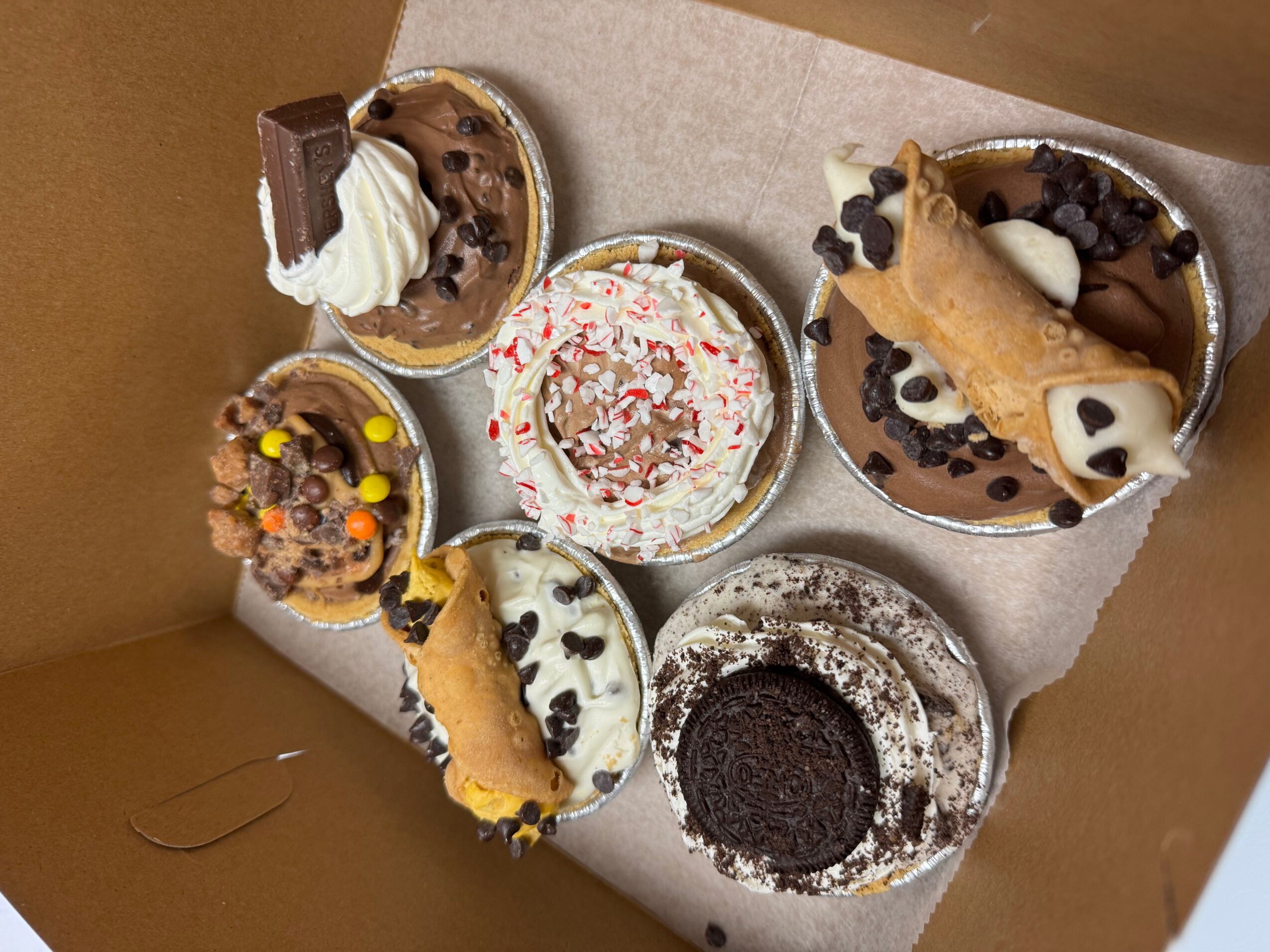 Six desserts in a box, including ice cream, cookies, and cake, with various toppings and decorations, on a beige surface.