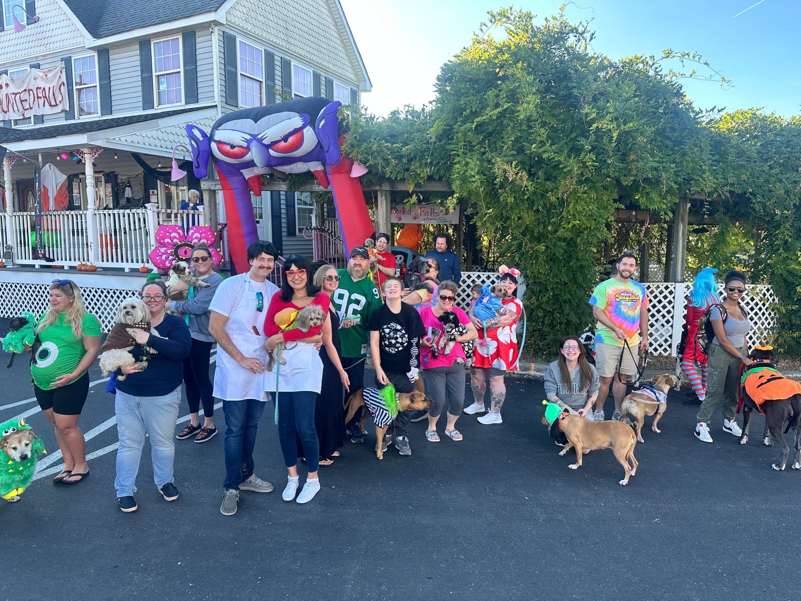 Group of people gathered outside a building with a colorful inflatable arch and trees, some holding balloons, with a dog present.
