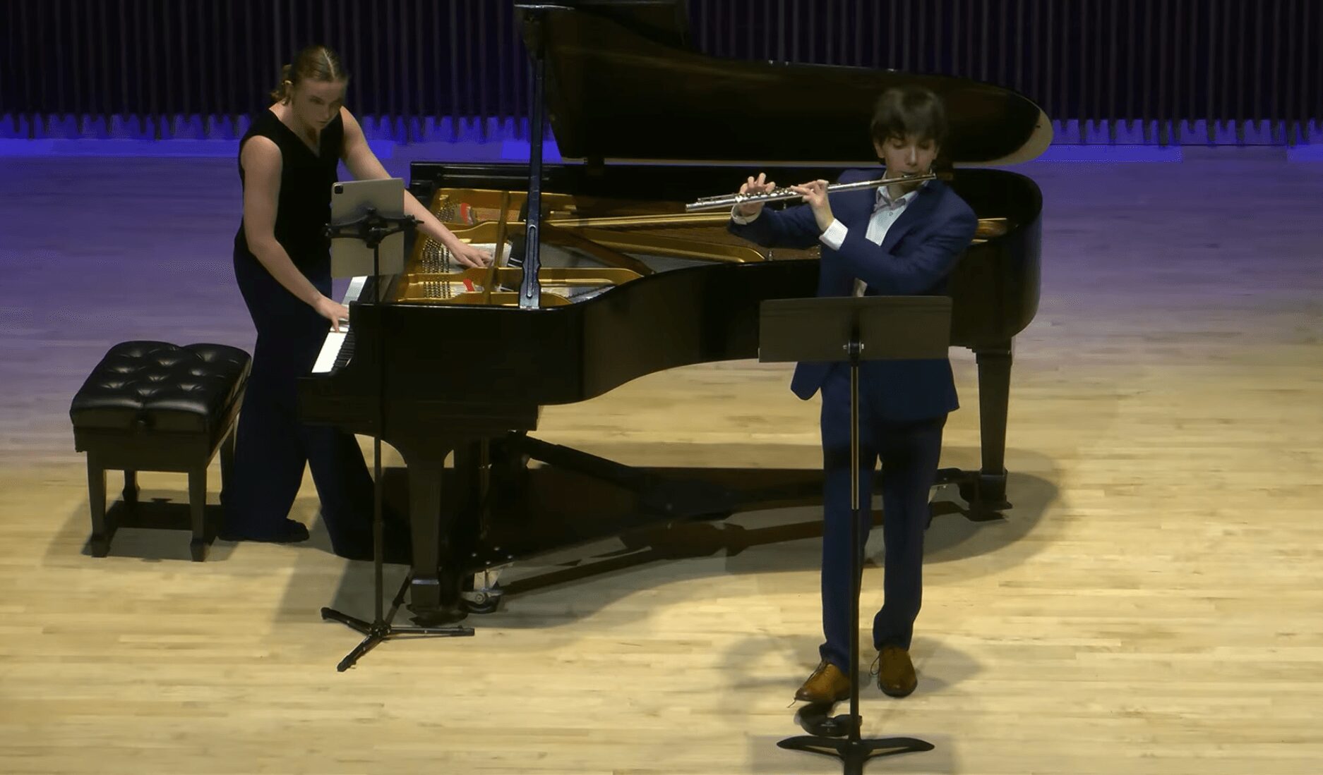 Two musicians perform on stage with a grand piano; one plays the piano, the other plays the flute.