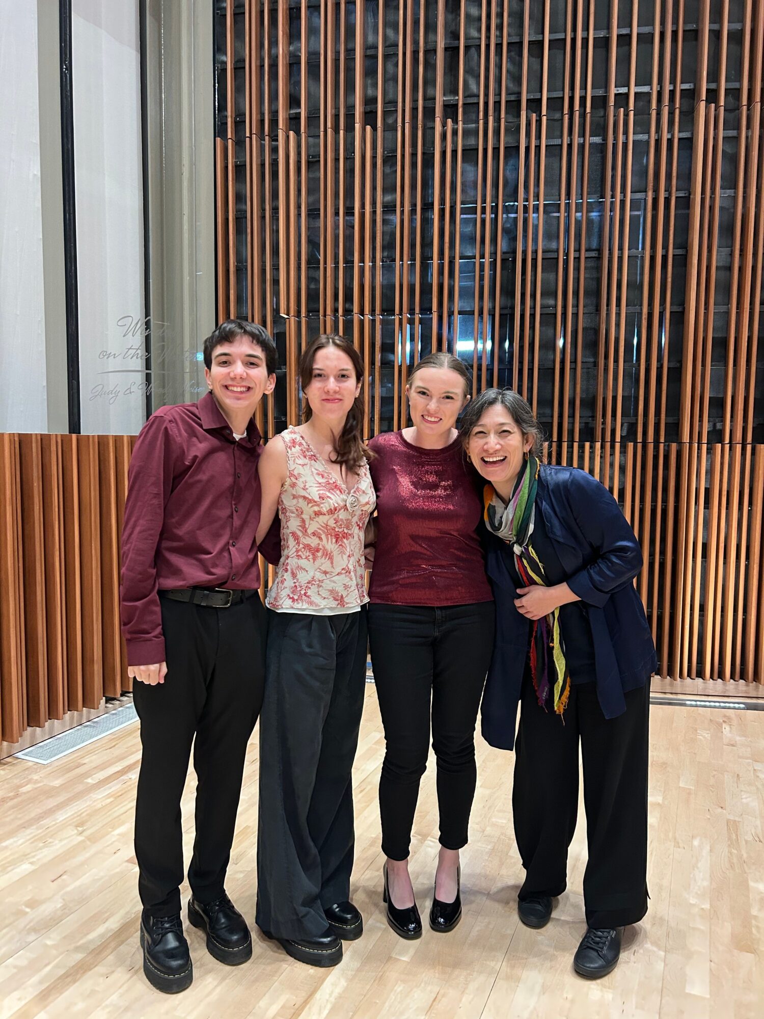 Four people standing indoors in front of a wooden slat wall, smiling and posing for the photo.