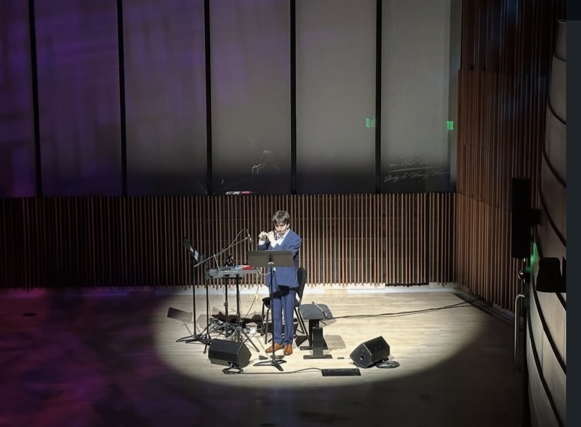 Person standing on stage with musical instruments, performing under spotlight in a large auditorium.