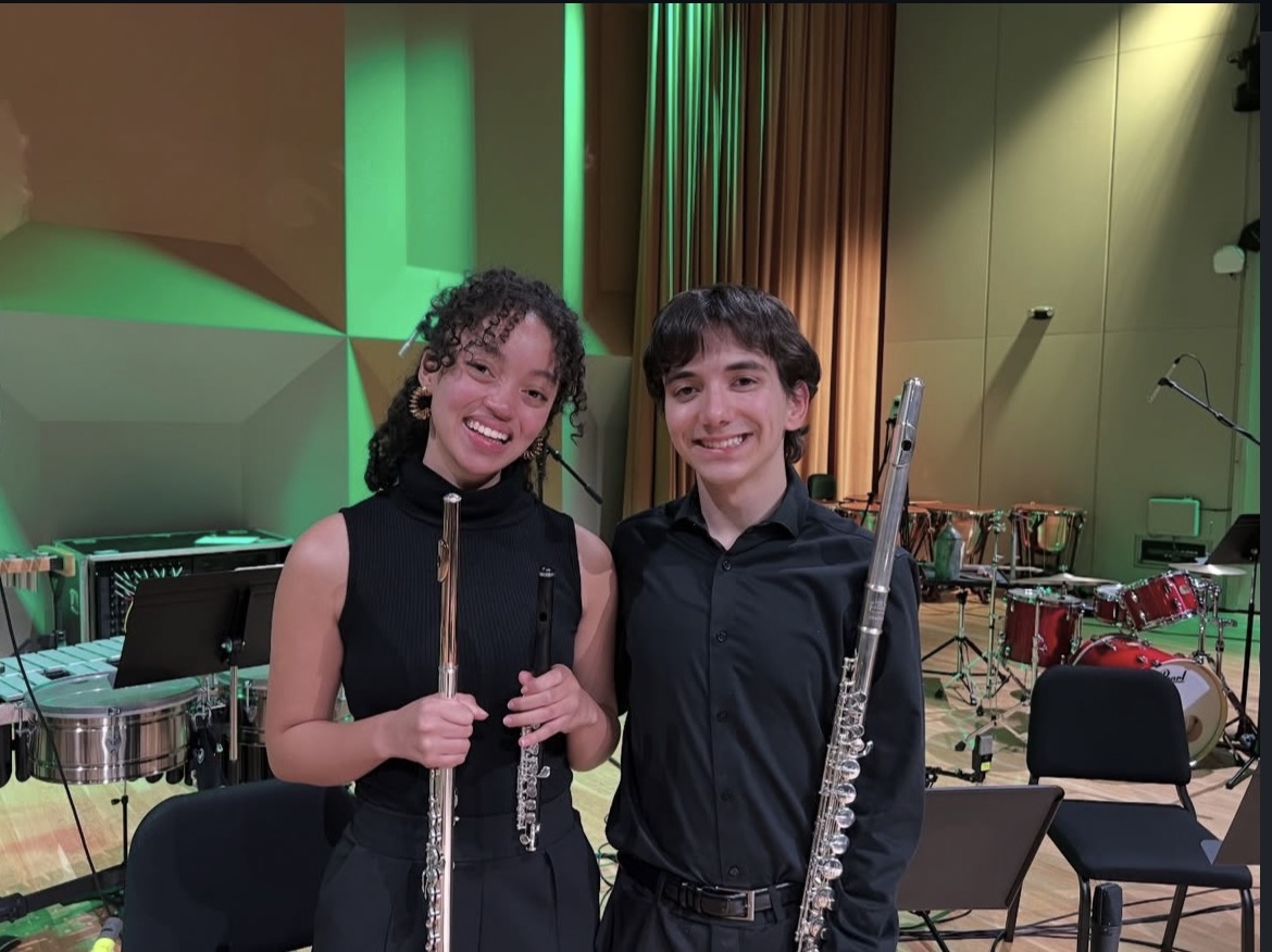 Two young people holding musical instruments, smiling, in a concert hall with stage and chairs in the background.