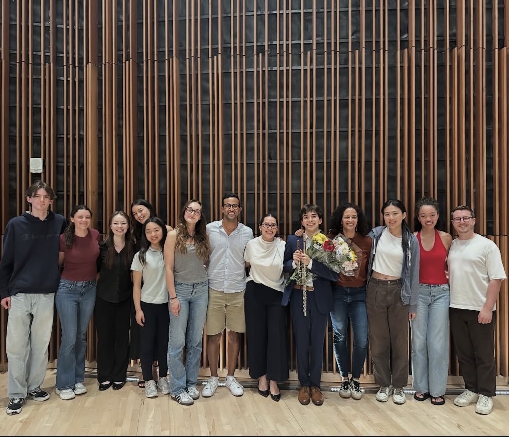 Group of people standing in front of a wooden wall, some holding flowers, smiling, indoors.