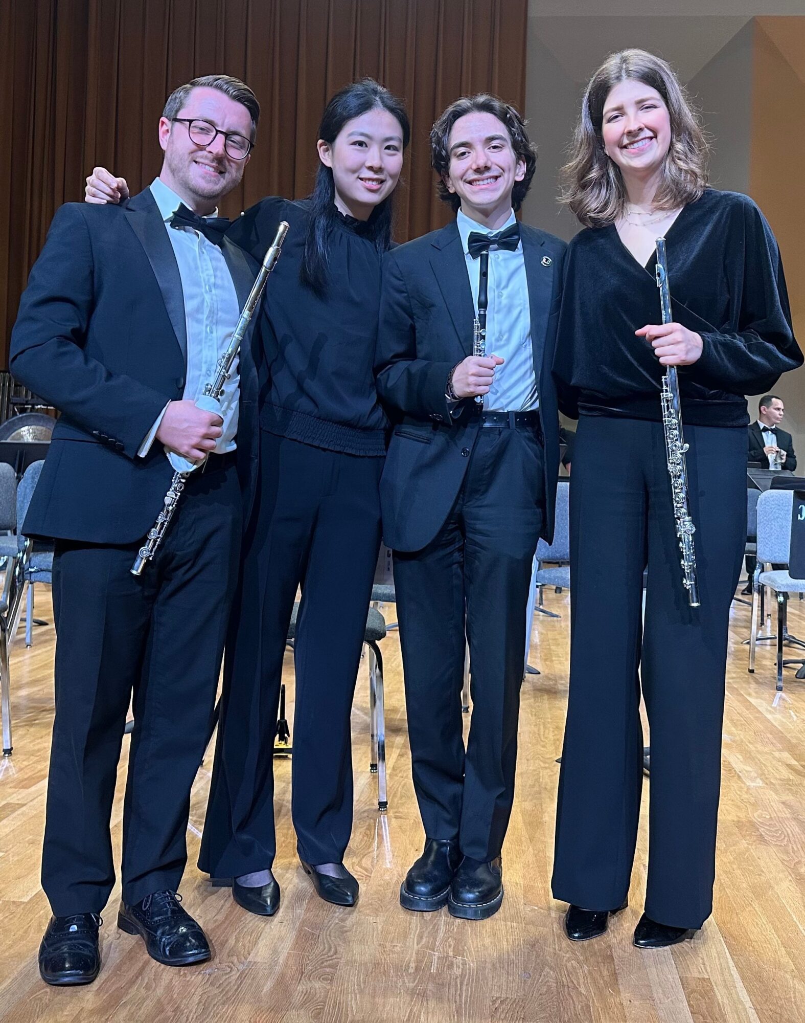 Four people standing together holding flutes, smiling, in a room with chairs and a wooden floor.