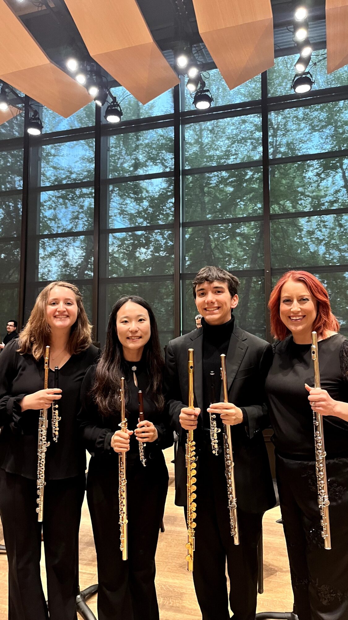 Four women holding flutes, standing indoors with large window and trees outside, smiling, dressed in black, concert setting.
