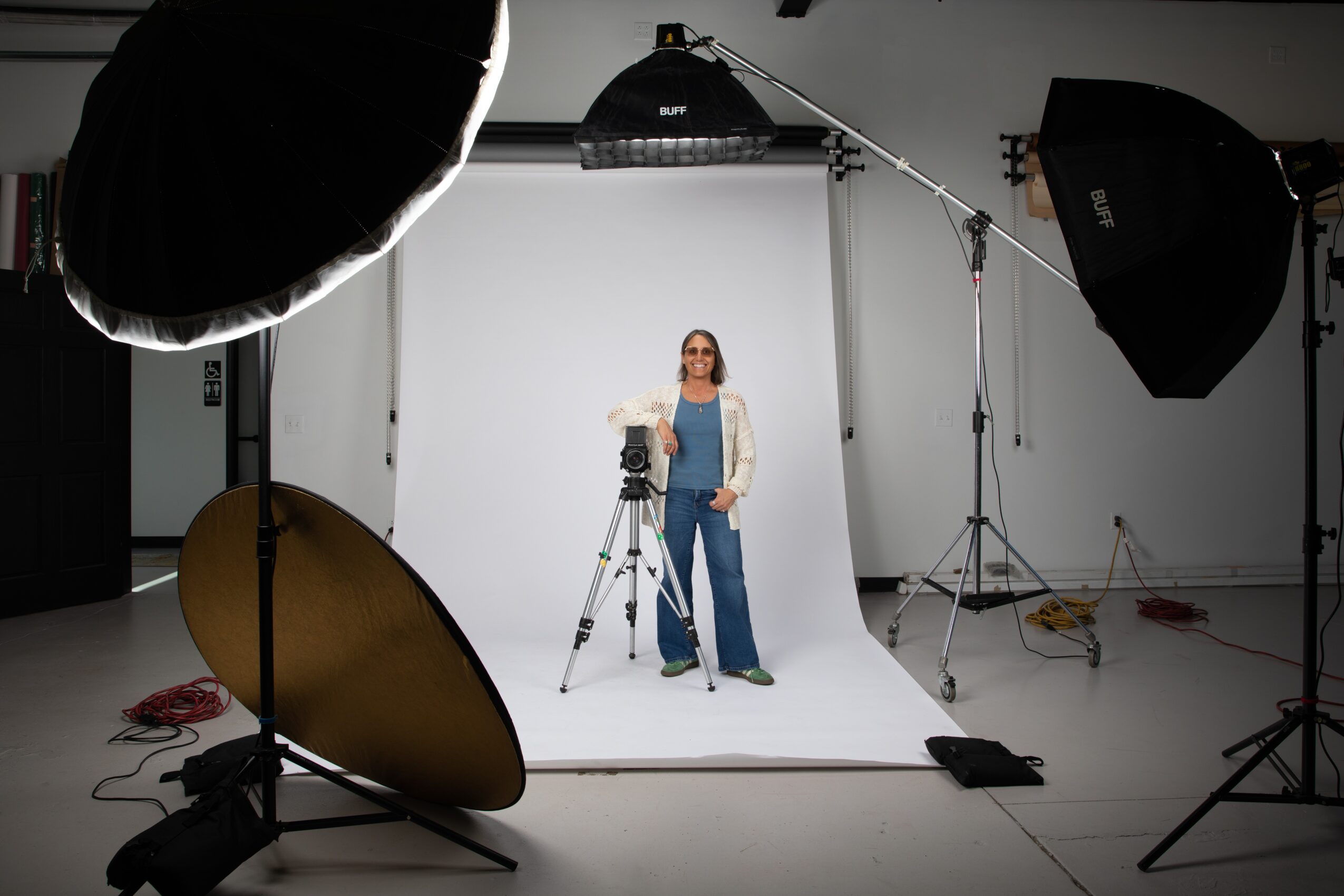Woman standing in a photography studio with lighting equipment and a white backdrop, camera on tripod, wearing blue overalls and white jacket.