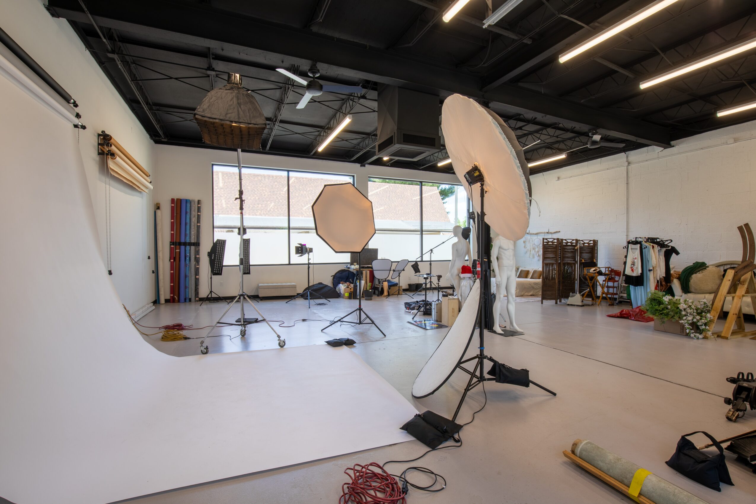 Photography studio with lighting equipment, large white backdrop, and various props, set up in a spacious room with large windows.