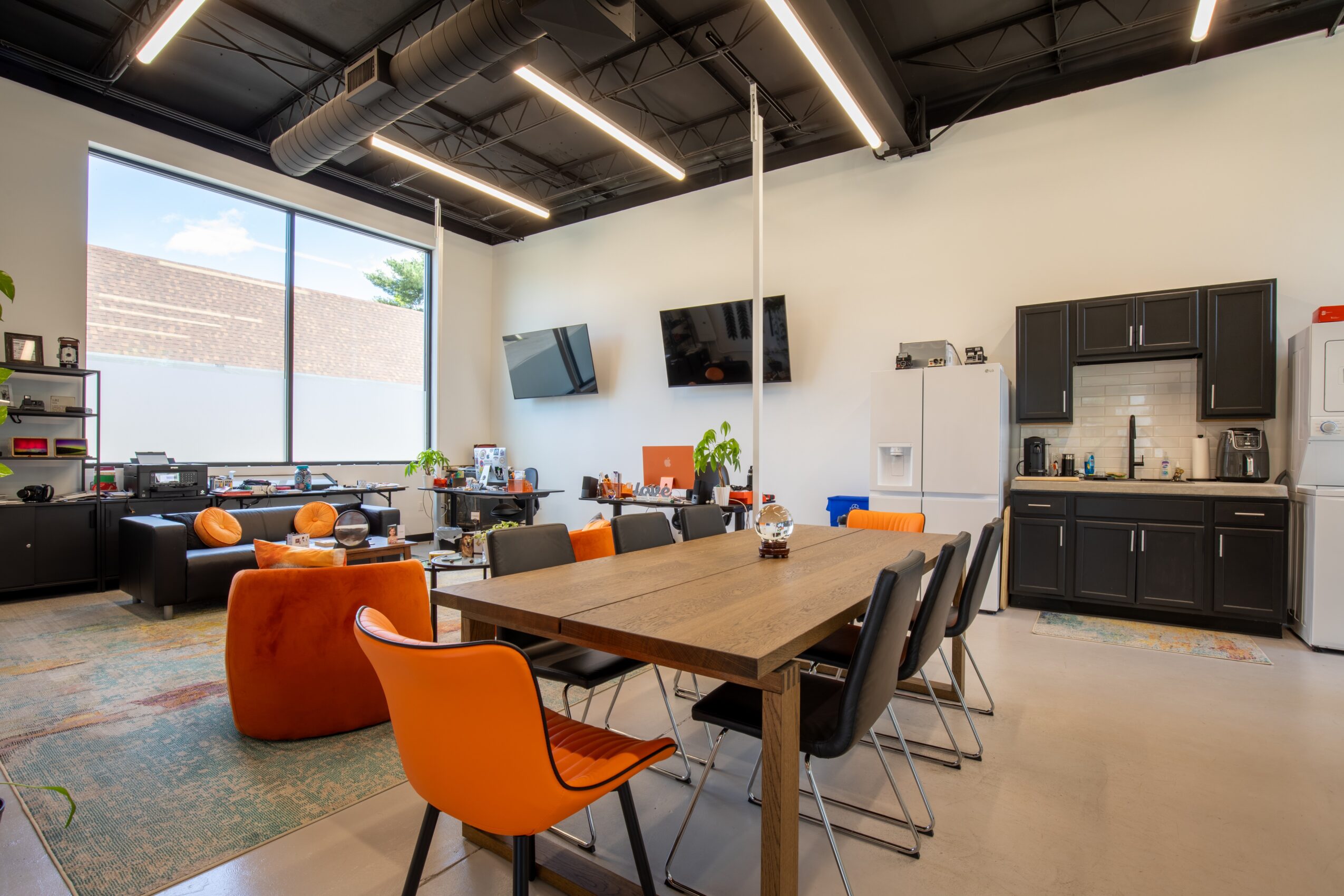Modern lounge with large window, tables, orange and black chairs, wall-mounted TVs, and a kitchenette area.