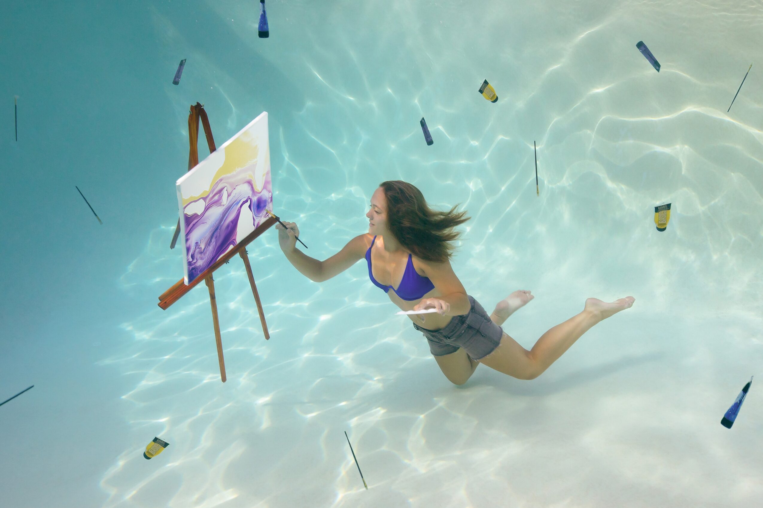 Woman in a blue swimsuit painting on an easel underwater in a pool, surrounded by floating objects.