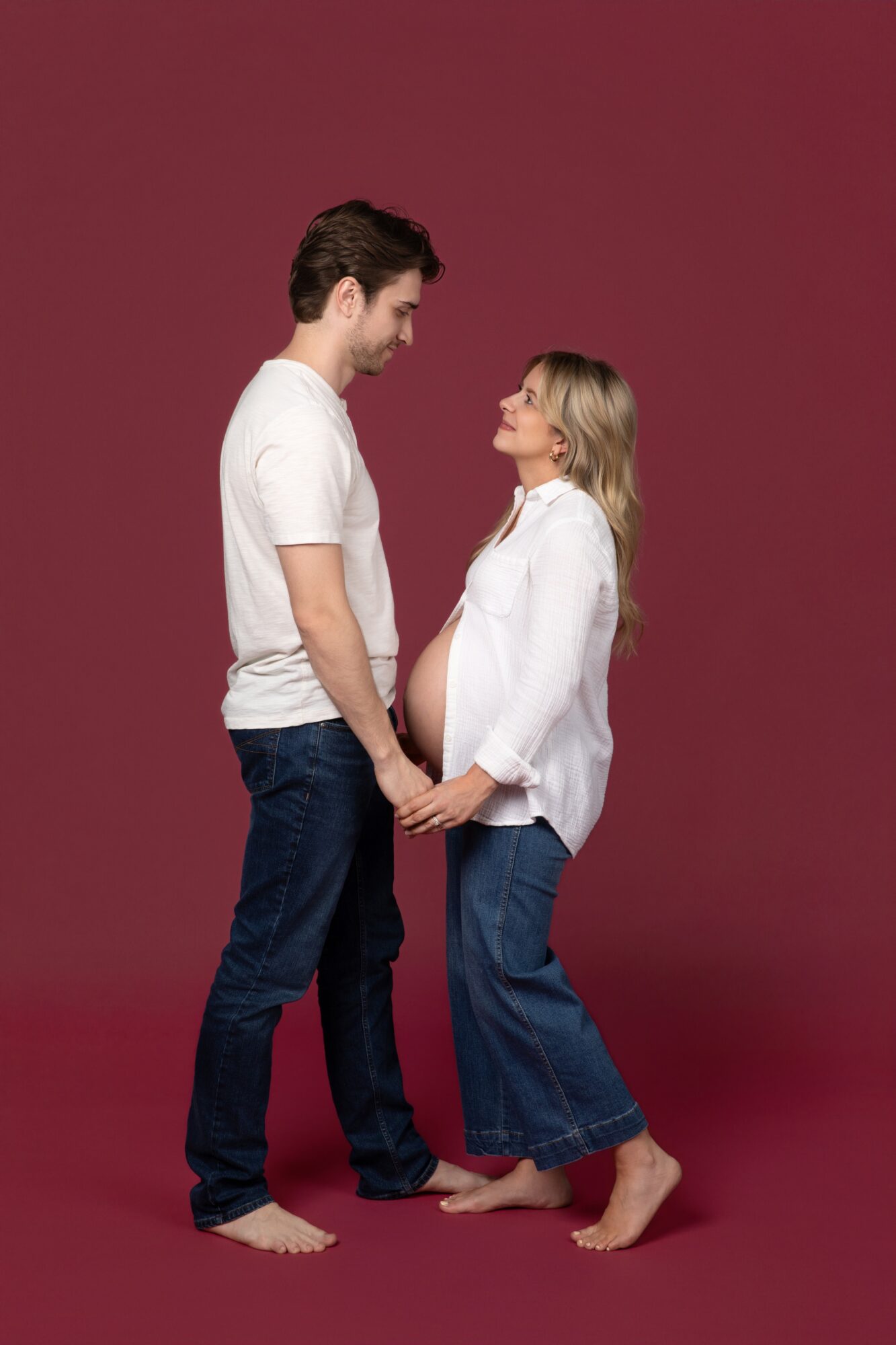 A pregnant woman and a man hold hands, facing each other, smiling, against a plain maroon background.