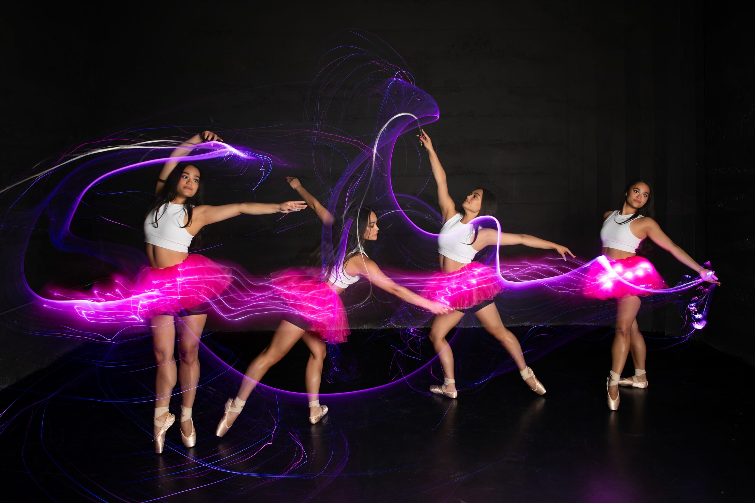 Four ballet dancers in pink tutus perform on stage with light trails around them.