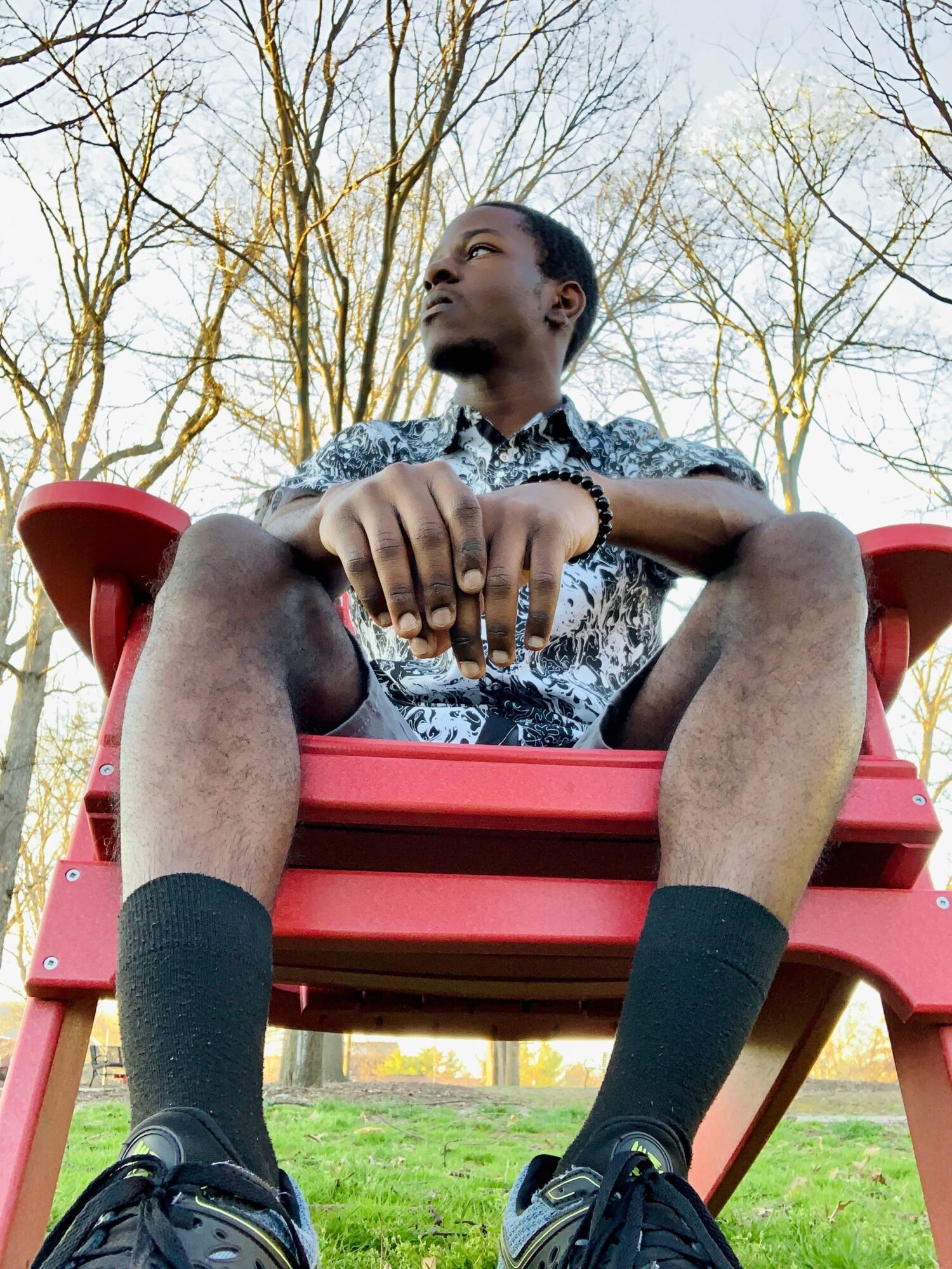 Young man sitting on a red outdoor chair with trees in the background, looking upward, wearing a patterned shirt and black socks.