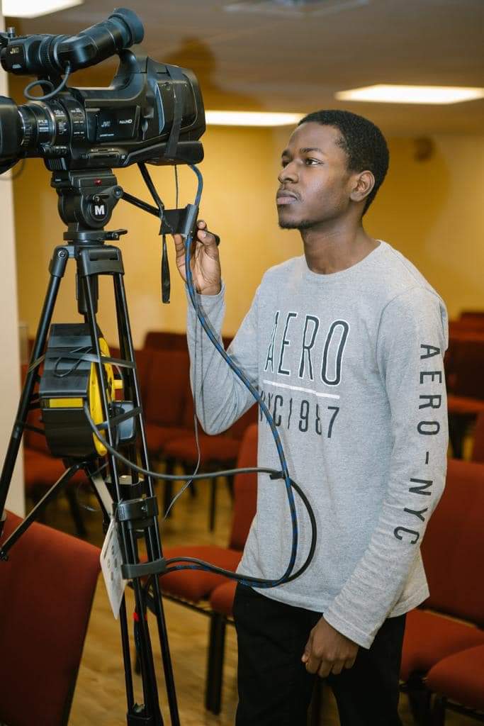 Young man operating a professional video camera indoors, with chairs and a yellow wall in the background.