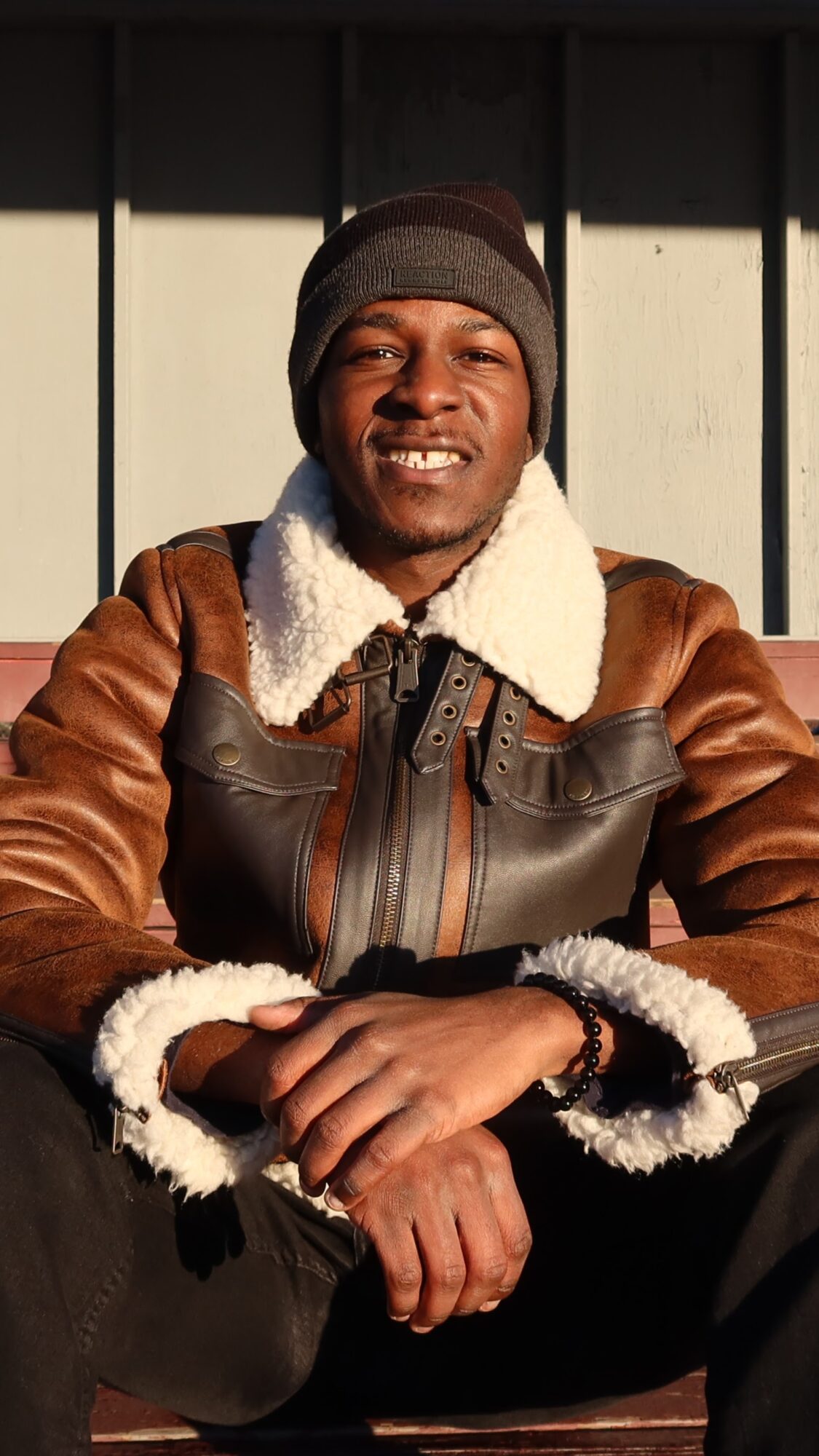Smiling man wearing a beanie and a brown leather jacket with a white collar, sitting outdoors in front of a wooden wall.