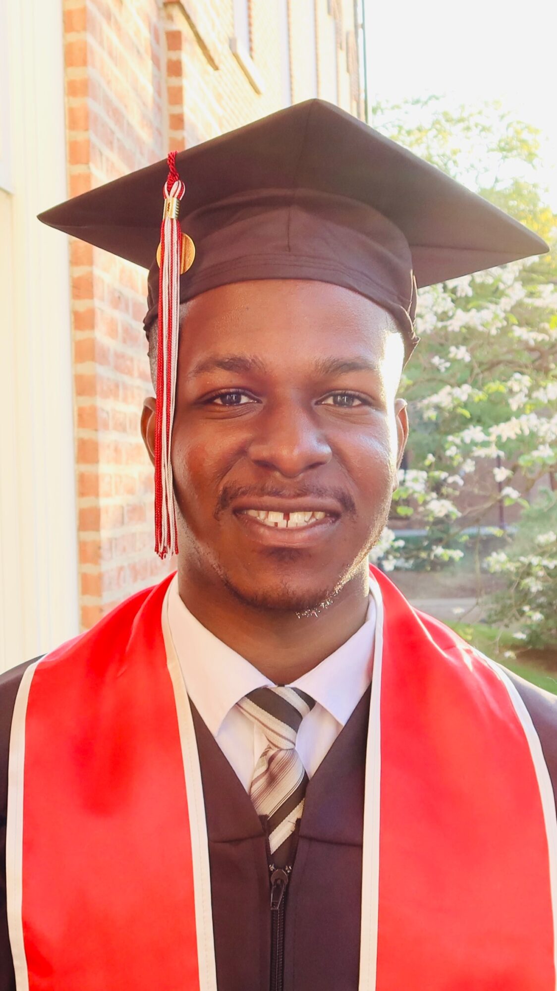 Young man in graduation cap and gown smiling outdoors with trees and building in background.