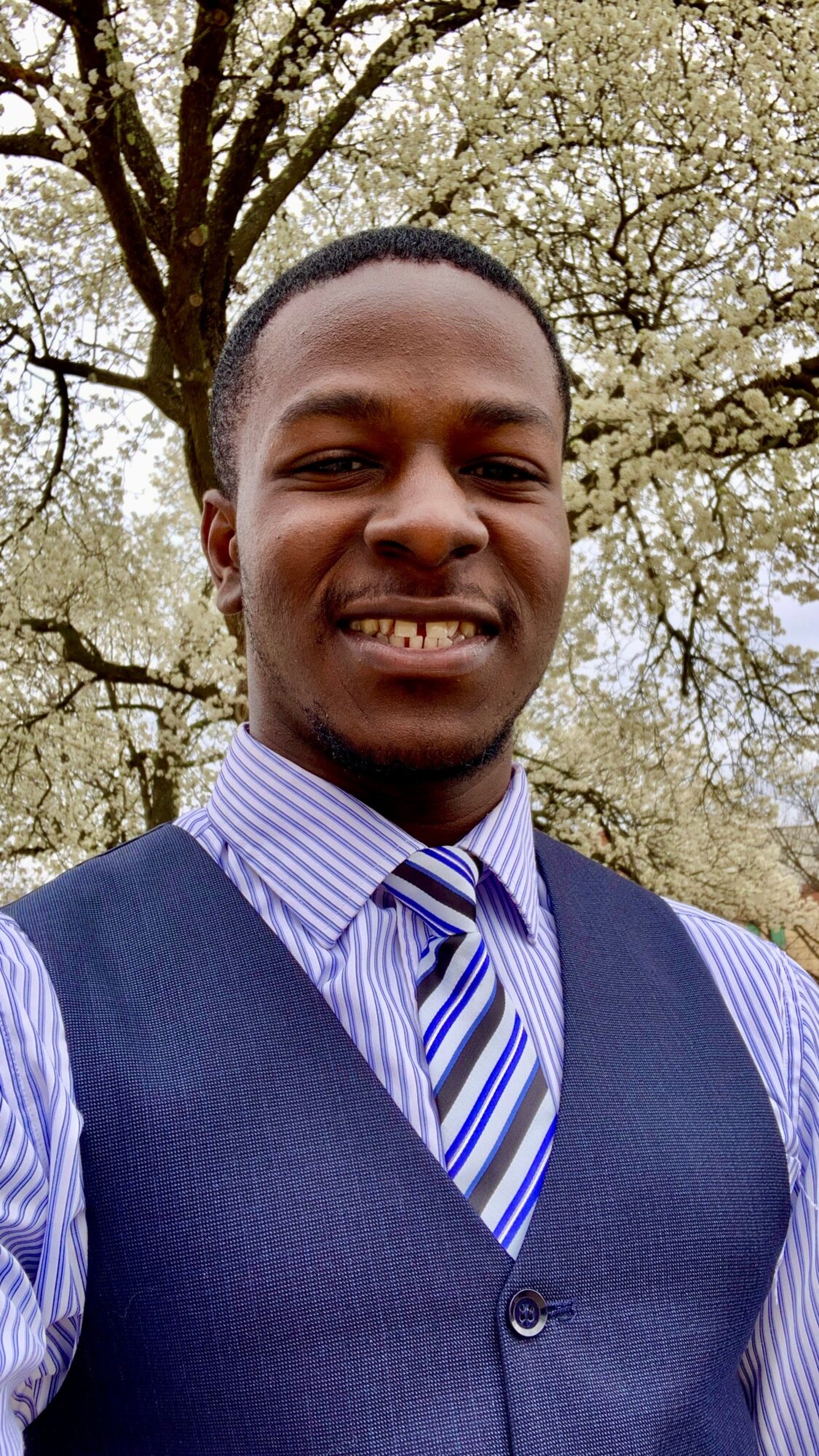 Young man in formal attire outdoors with trees in background, smiling, wearing a striped tie and vest.