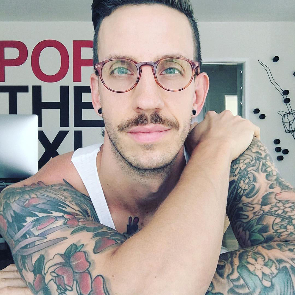 Man with glasses, mustache, tattoos on arms, wearing a white tank top, resting his arm on his shoulder, in a room with wall art.