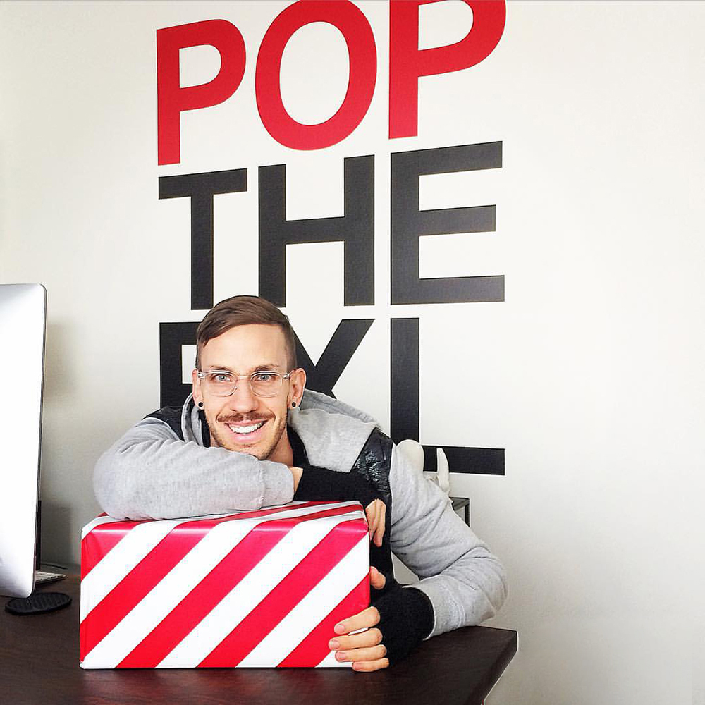 Man with glasses smiling, sitting at desk with a red and white striped box, behind a wall with large red and black text.