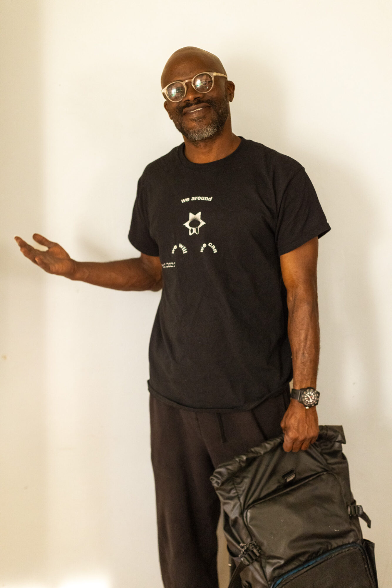 Smiling man with glasses and beard, wearing a black t-shirt, holding a backpack, standing against a plain white background.