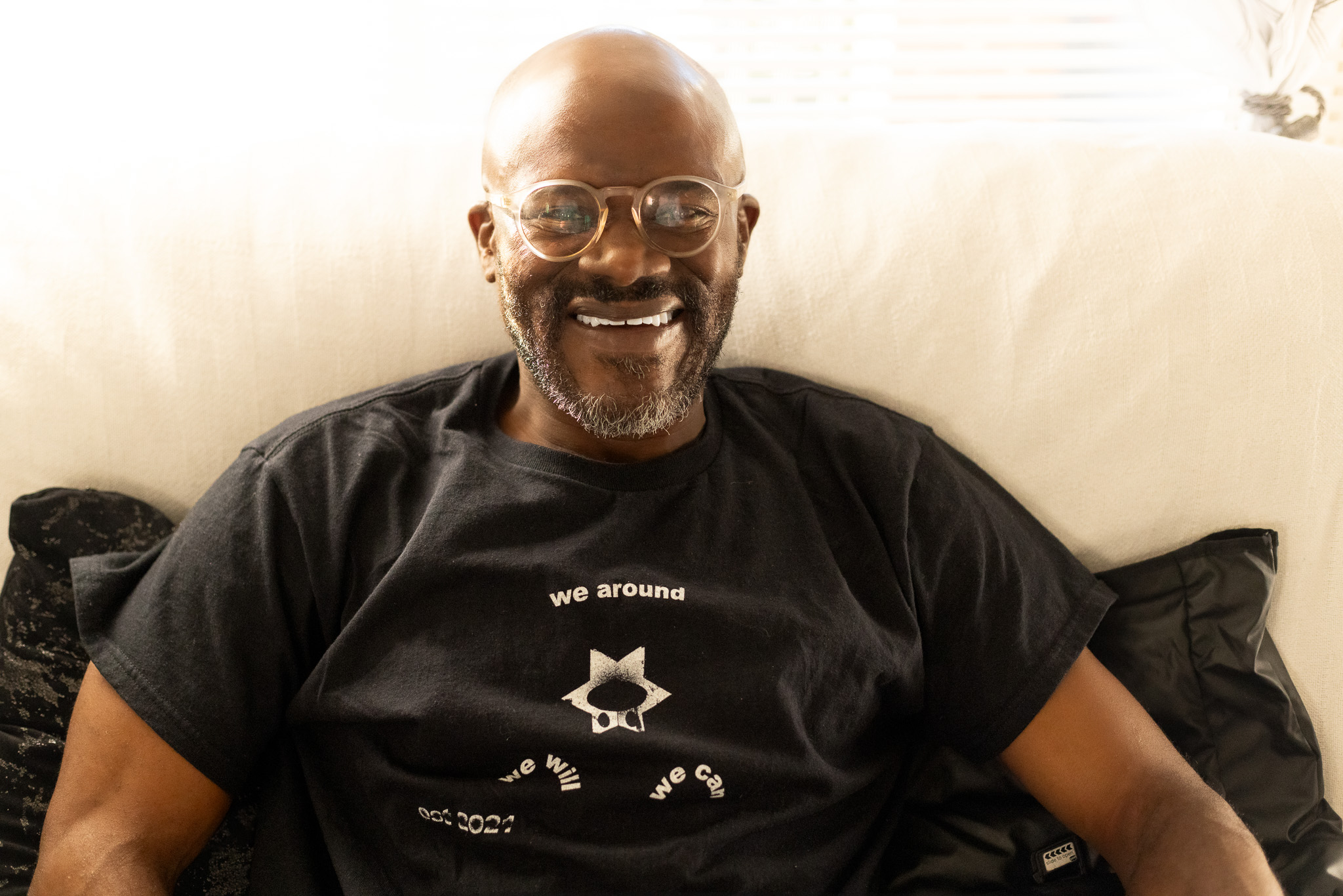 Smiling man with glasses and a beard sitting on a sofa, wearing a black T-shirt with text and graphics.