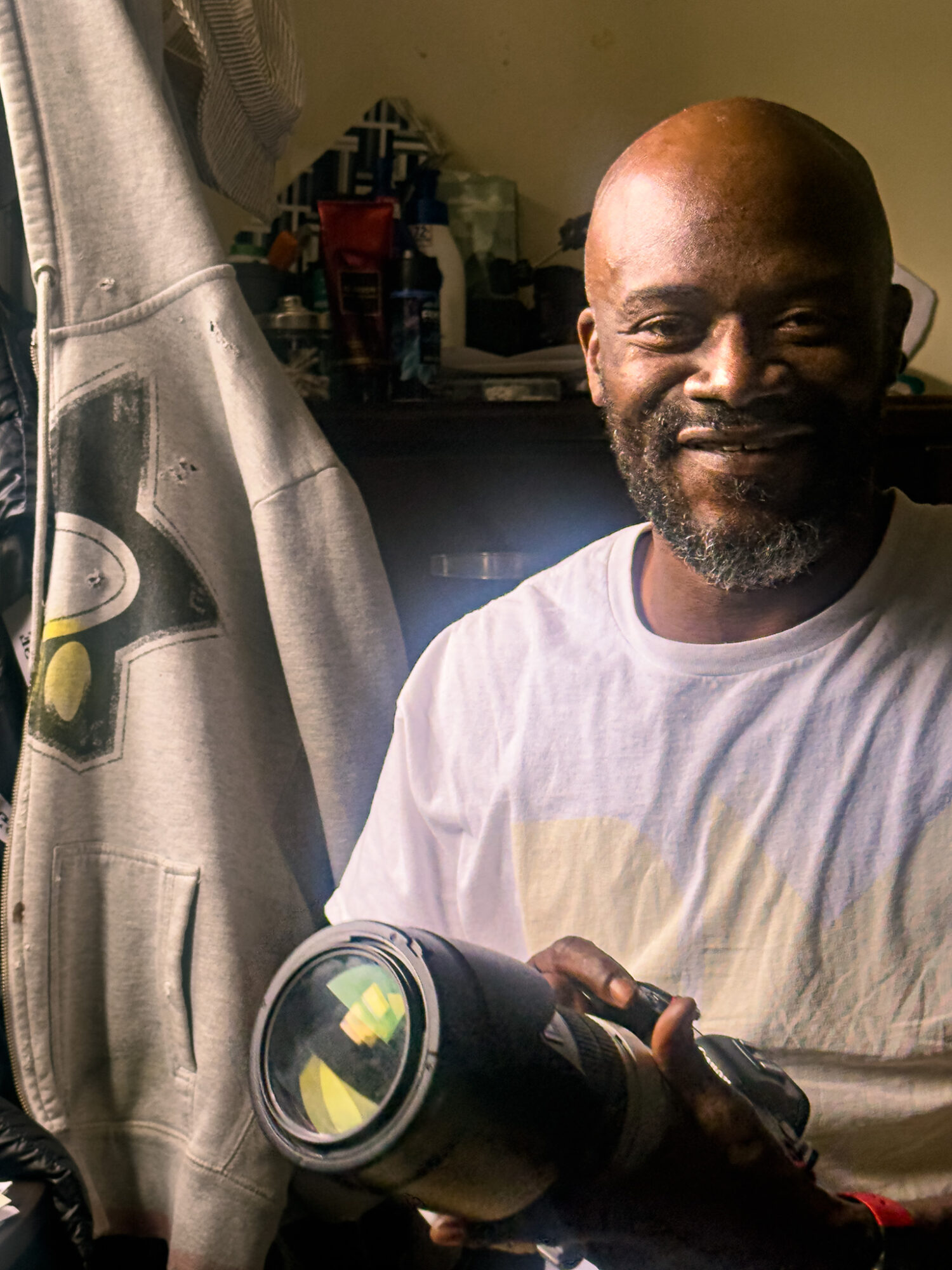 Man with a beard holding a camera, smiling, in a room with a curtain and various objects in the background.