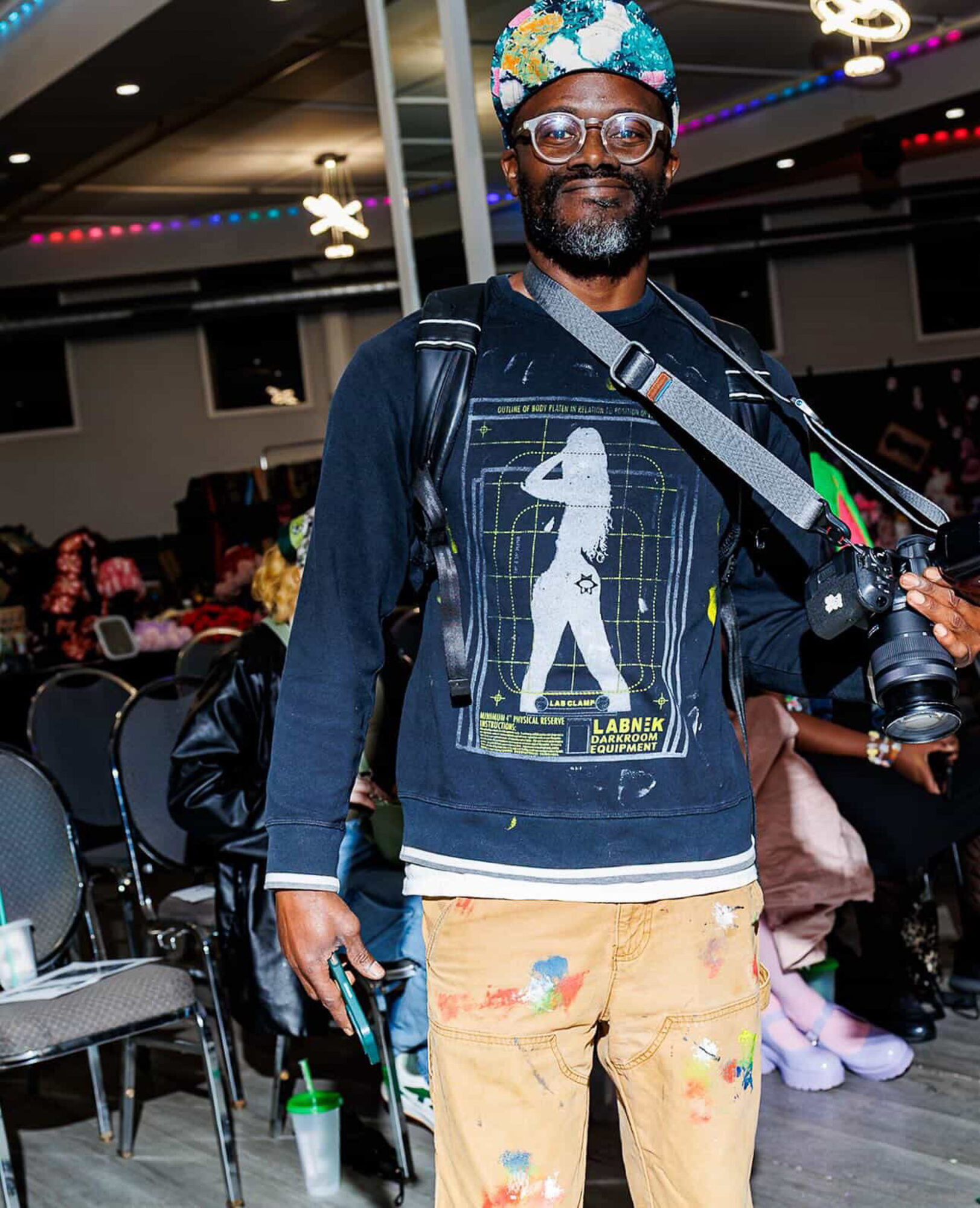 Man with glasses, colorful headwrap, and backpack standing indoors, smiling, with chairs and people in the background.