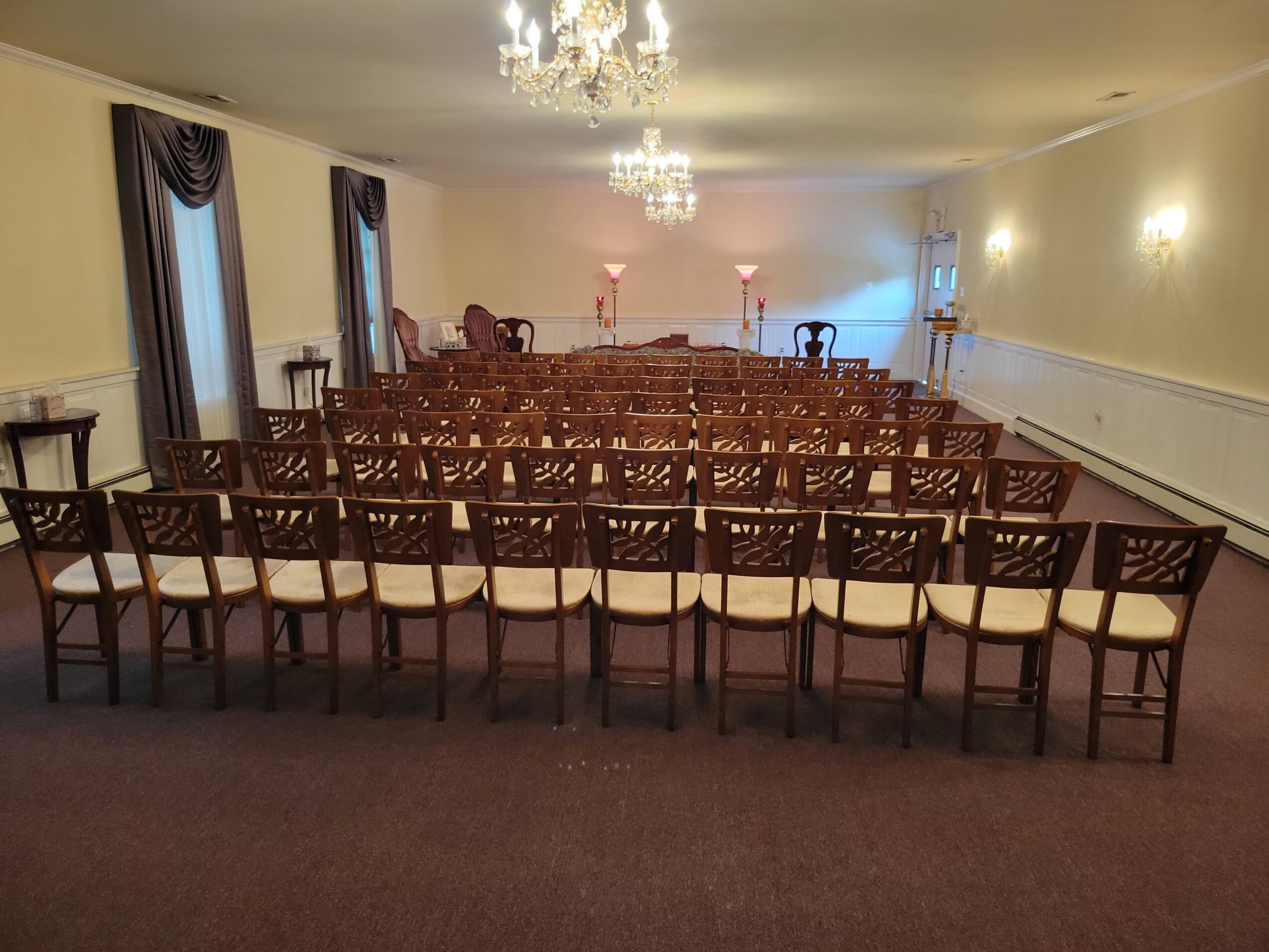 Empty room with rows of chairs facing a wall with two lamps and a chandelier, curtains on windows, and a carpeted floor.