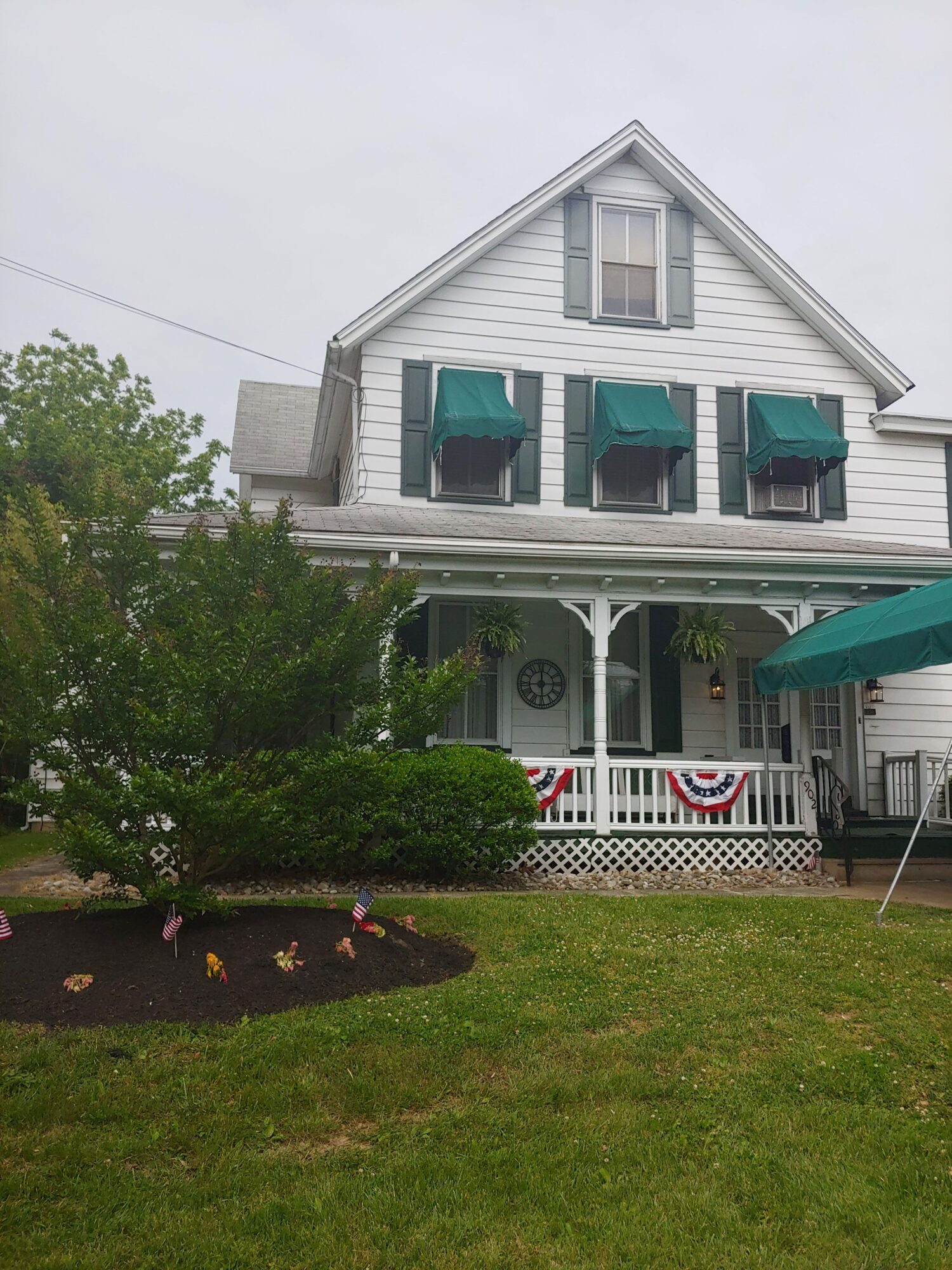 Two-story house with white siding, green shutters, and a front porch, surrounded by a lawn and trees.