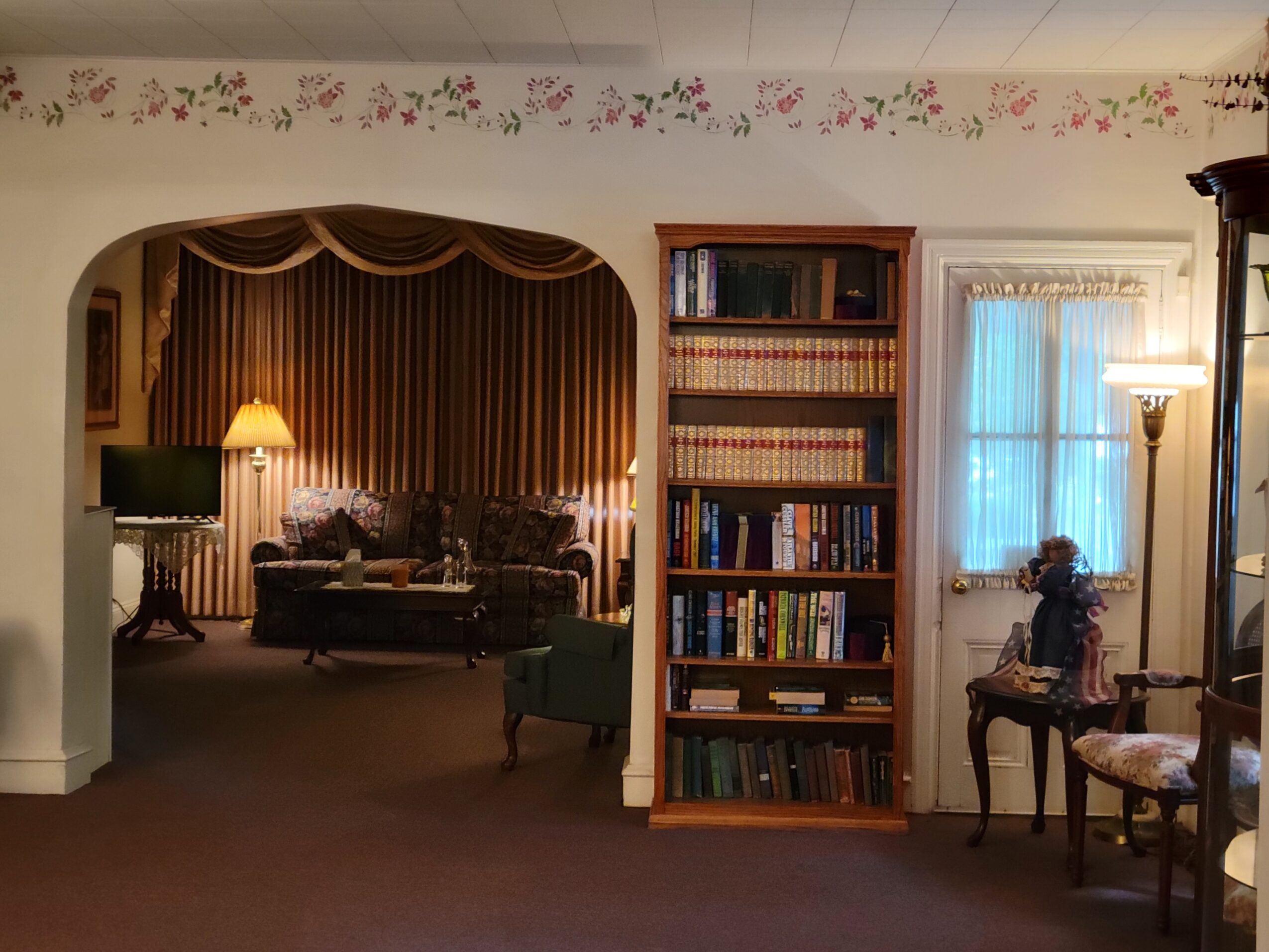 Living room with bookshelf, window, and seating area, decorated with floral border on ceiling, warm lighting, and curtains.