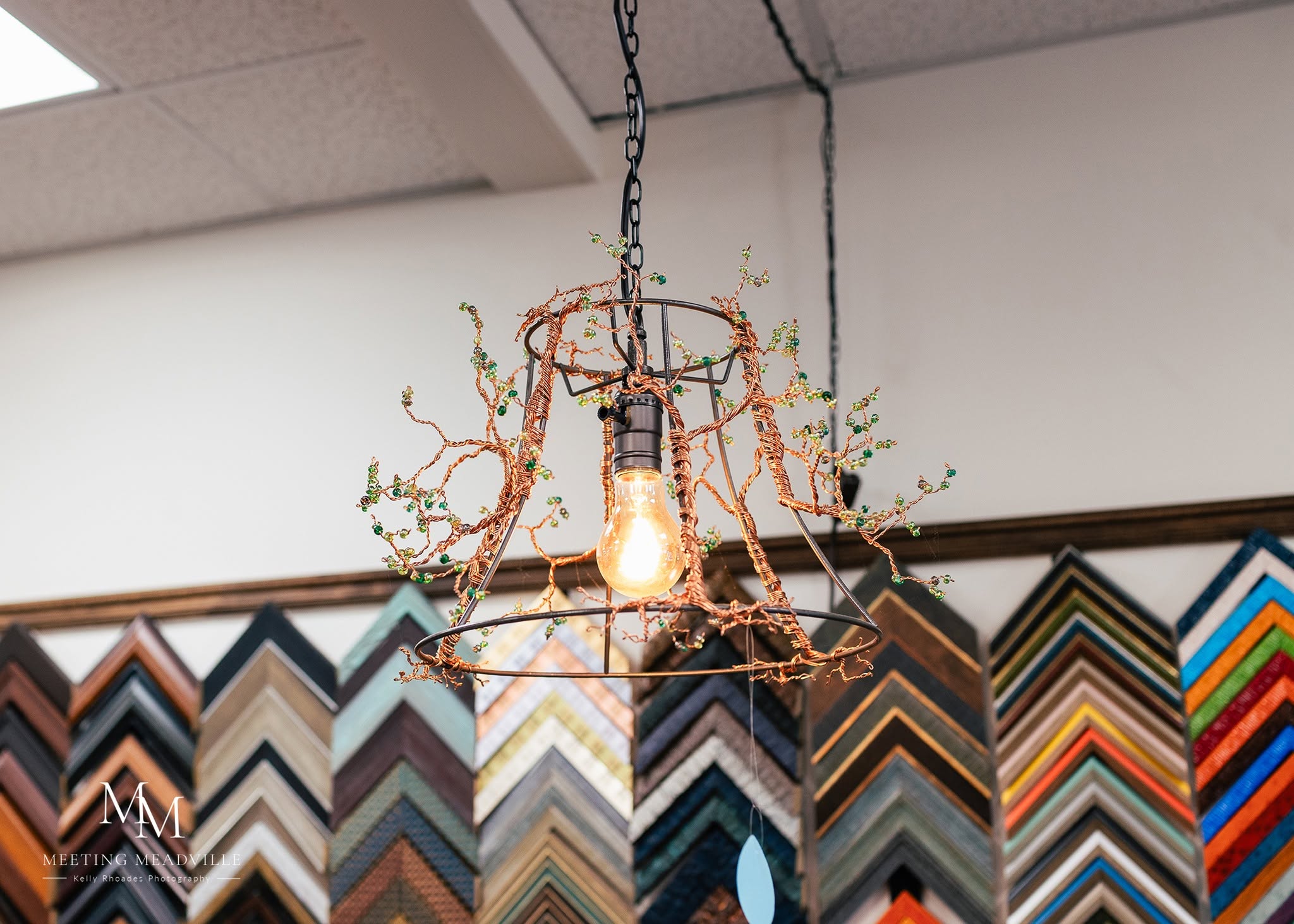 Light fixture with exposed bulb hanging from ceiling, surrounded by decorative branches, against a wall with colorful framing samples.