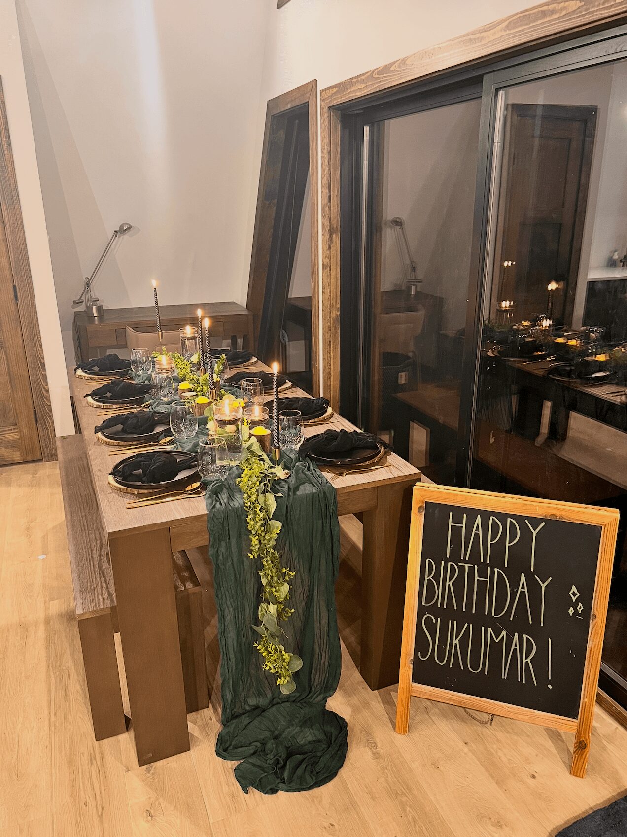 Party table with black plates, glasses, and a green floral centerpiece, next to a window with a birthday sign for Sukumar.