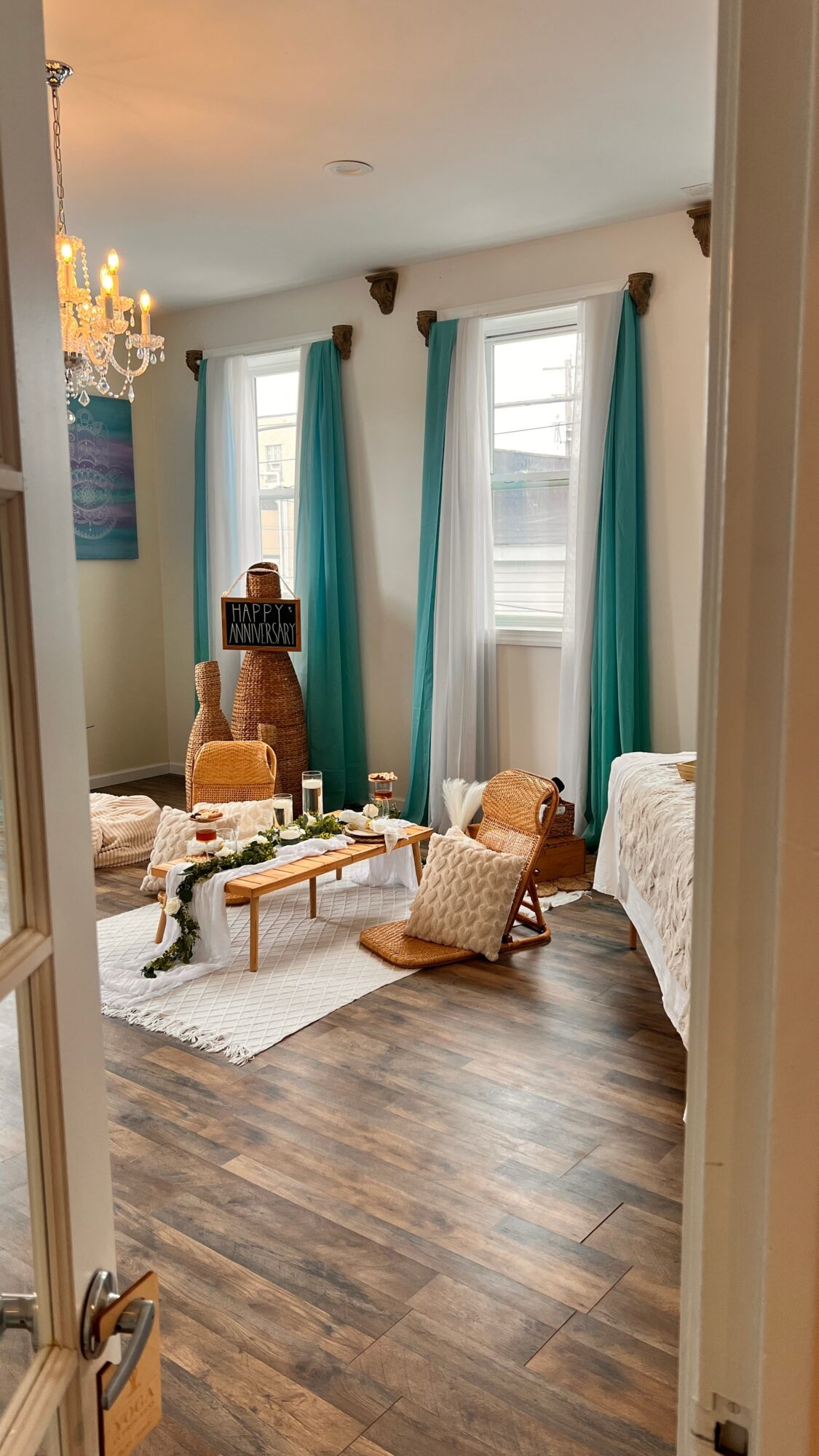 Living room with large windows, teal curtains, a chandelier, and furniture including a sofa, armchair, and coffee table.