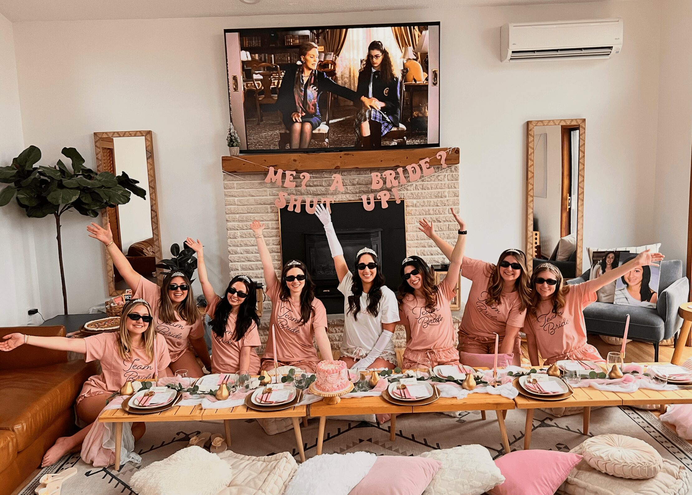 Group of women celebrating at a table with food, wearing matching pink outfits and sunglasses, in a decorated room.