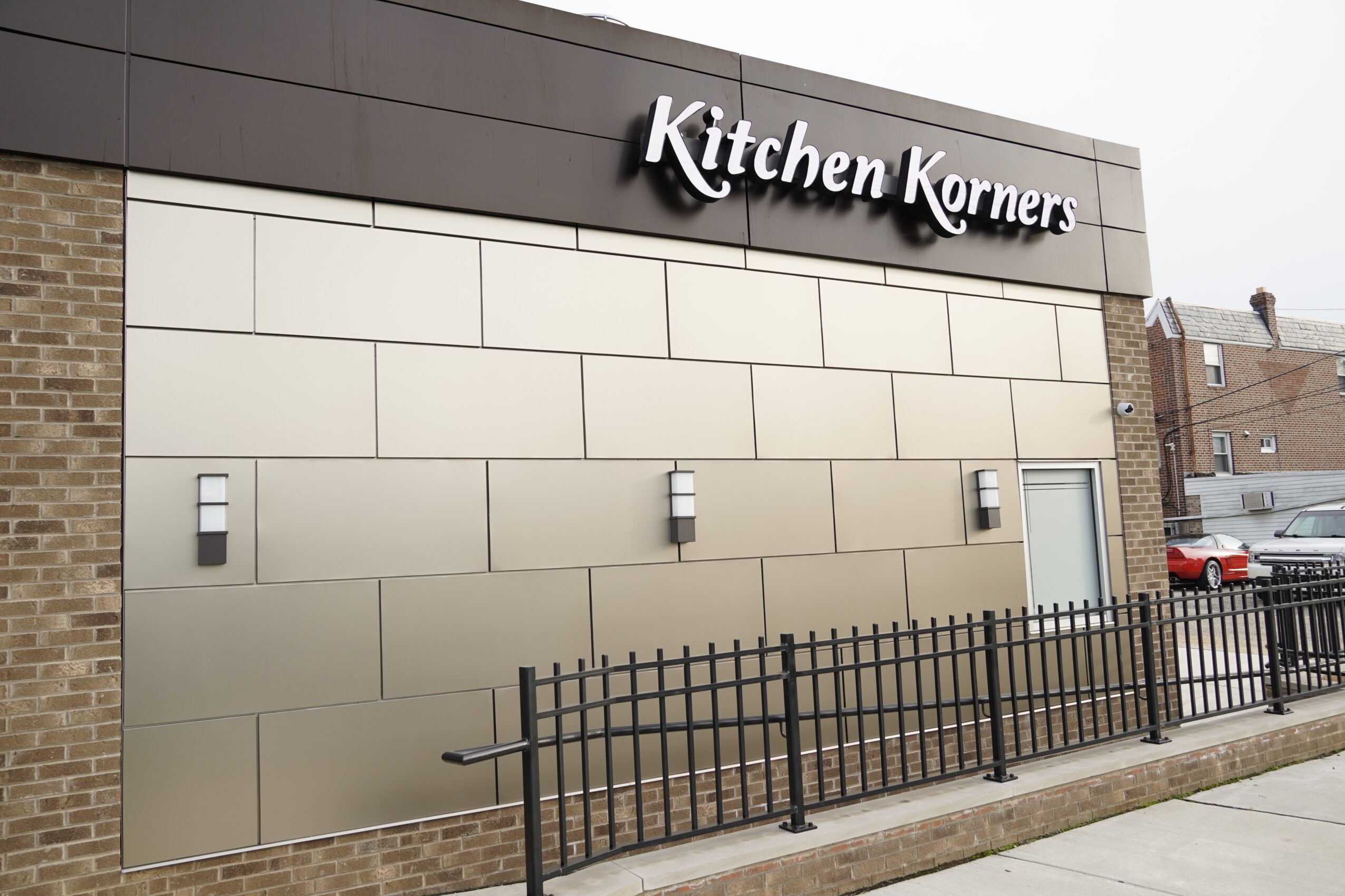 Exterior of a building with a sign reading 'Kitchen Kornerrs' and a black fence in front.