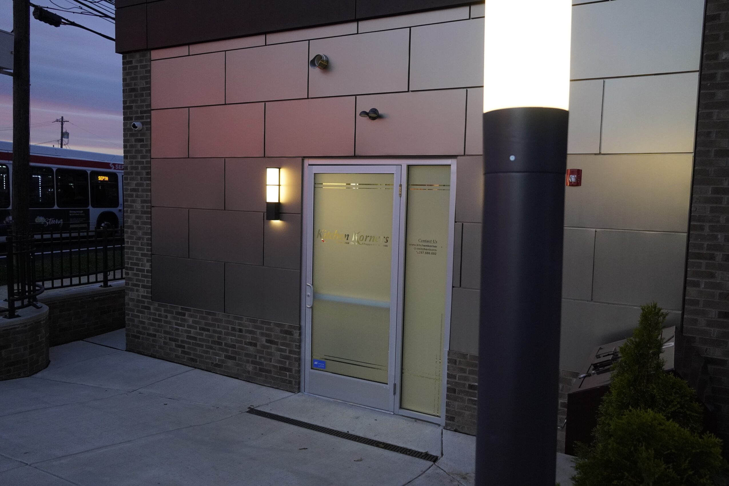 Exterior entrance with glass door, wall-mounted lights, and a tall illuminated column, with a sidewalk and street visible nearby.
