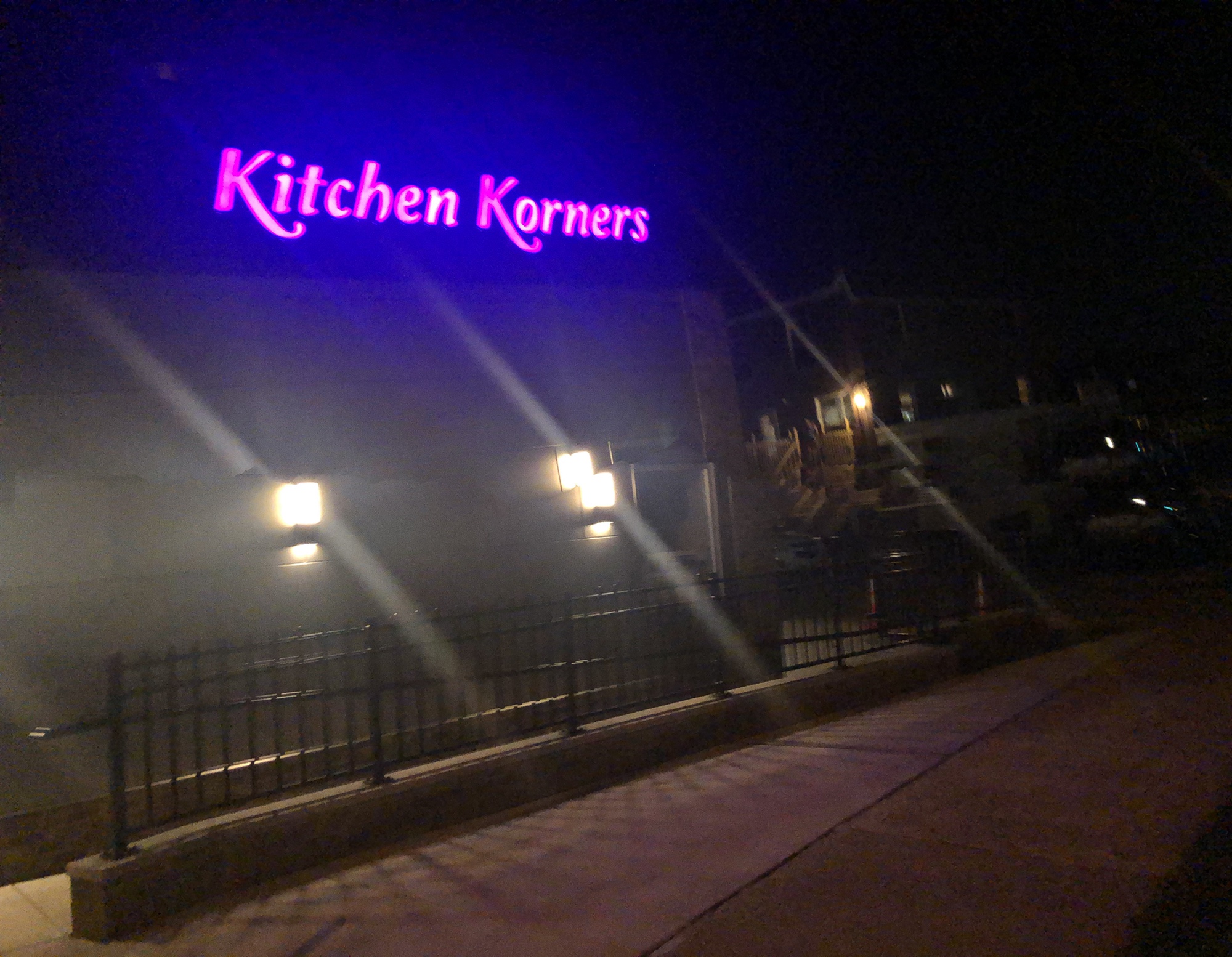 Neon sign reading 'Kitchen Korners' above a dark building, with a sidewalk and fence in front, at night.